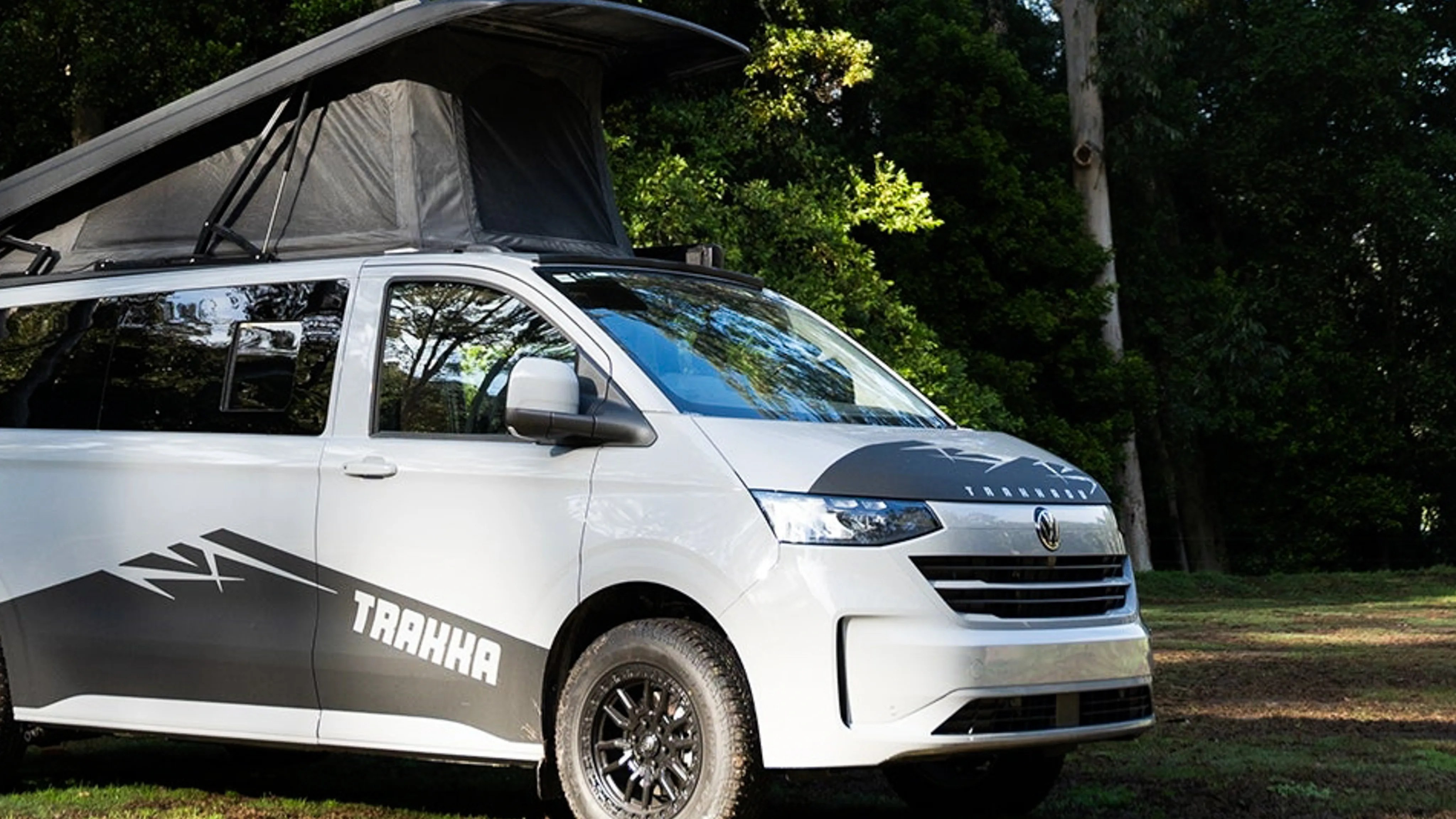 White Volkswagen camper van with black graphics and pop-up roof tent parked on grass near trees.