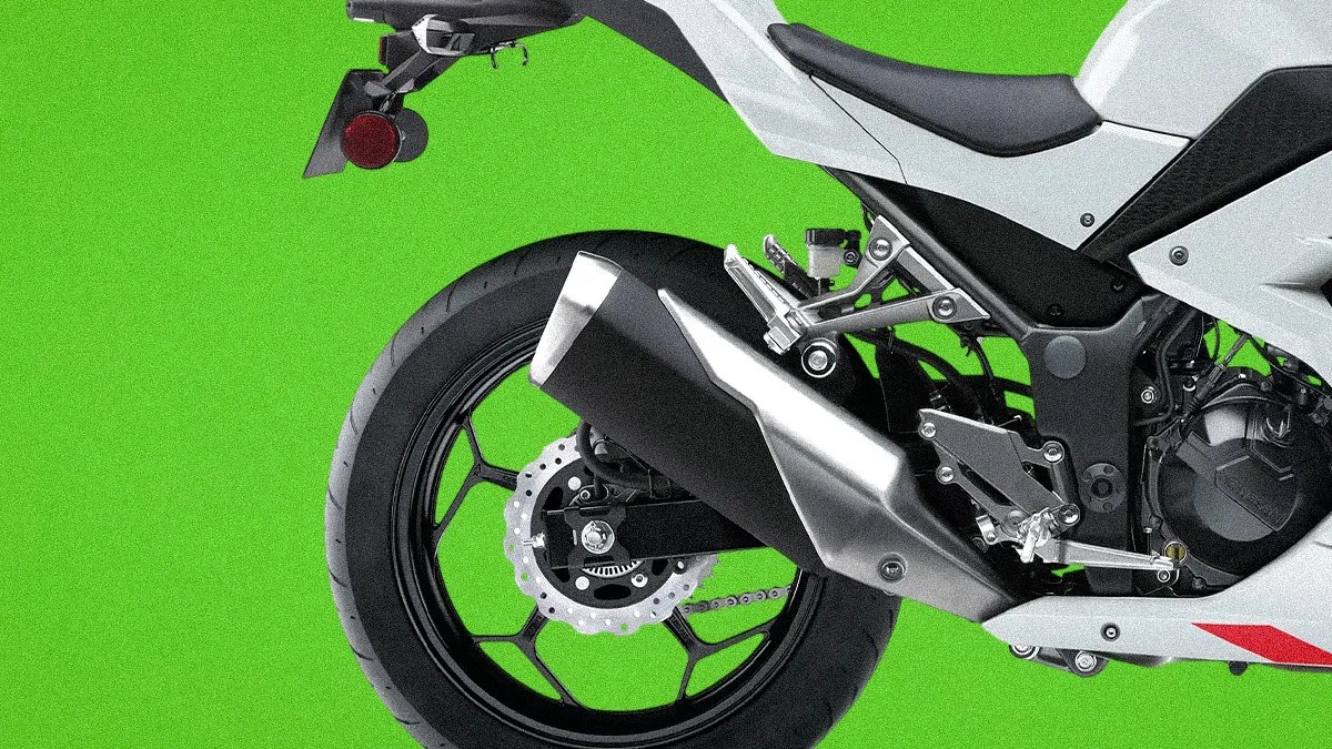 Rear section of a white motorcycle showing black rear tire, silver exhaust, and foot pegs against a green background.