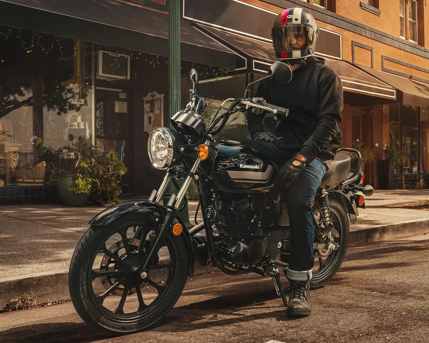 Person wearing a black hoodie, jeans, gloves, and a helmet sitting on a black Kawasaki motorcycle on a street.
