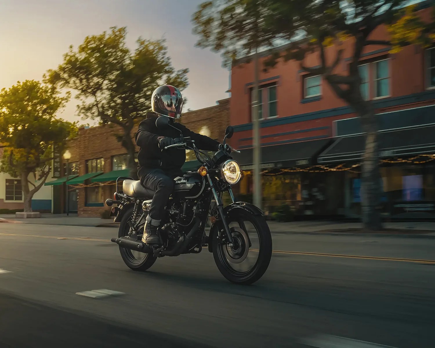 Person wearing black riding gear and helmet riding a black Kawasaki motorcycle on a street at sunset.