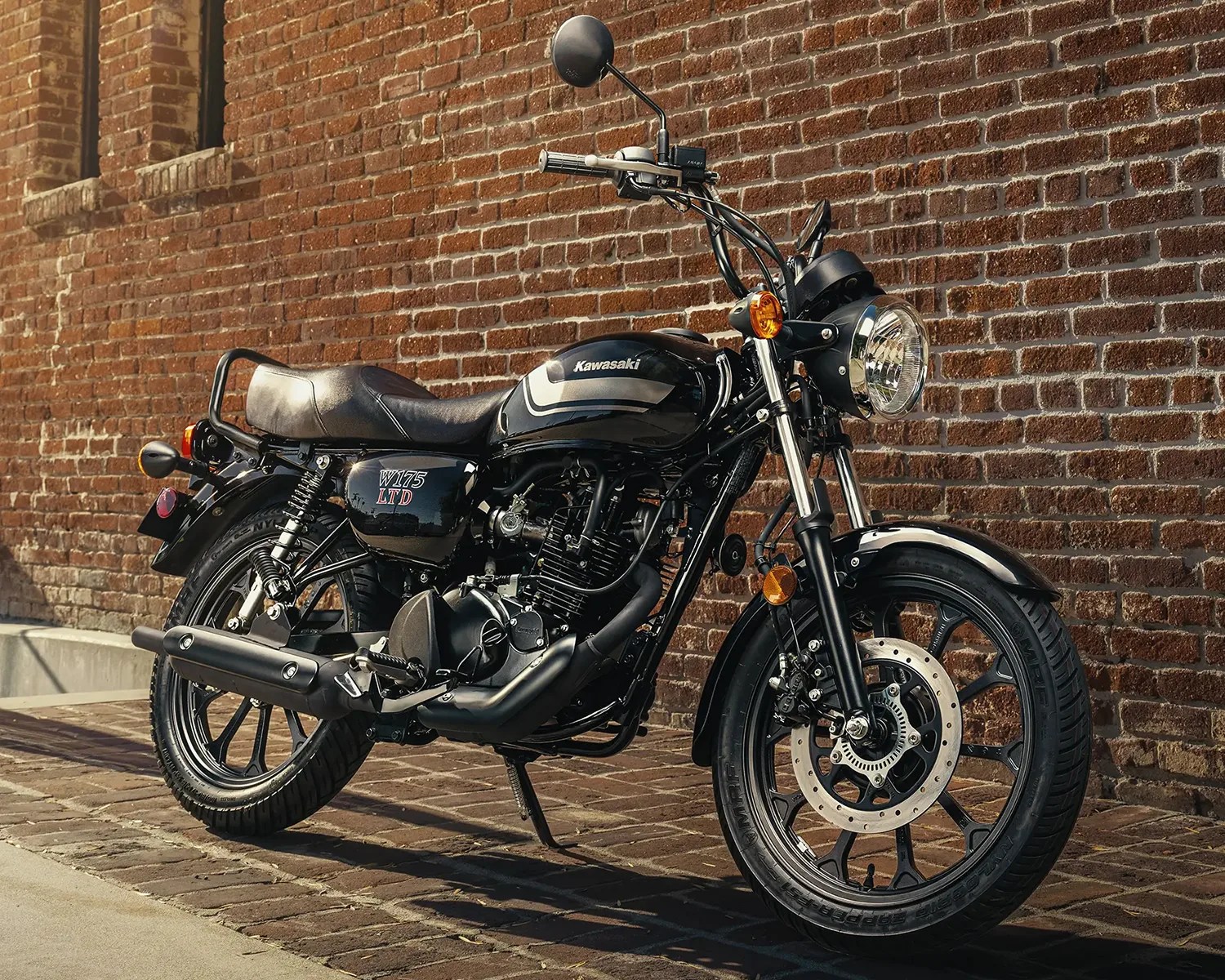 Black Kawasaki W175 LTD motorcycle parked on a brick pavement against a brick wall.