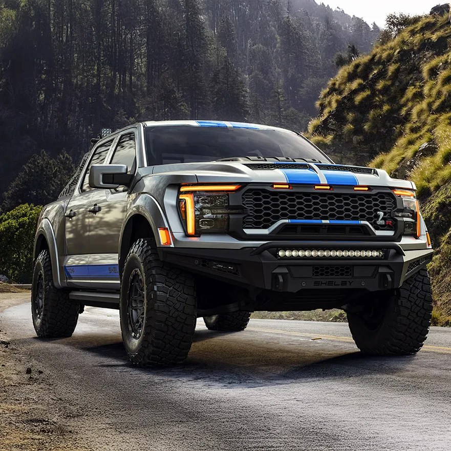 Silver and blue Shelby pickup truck with large off-road tires parked on a mountain road.