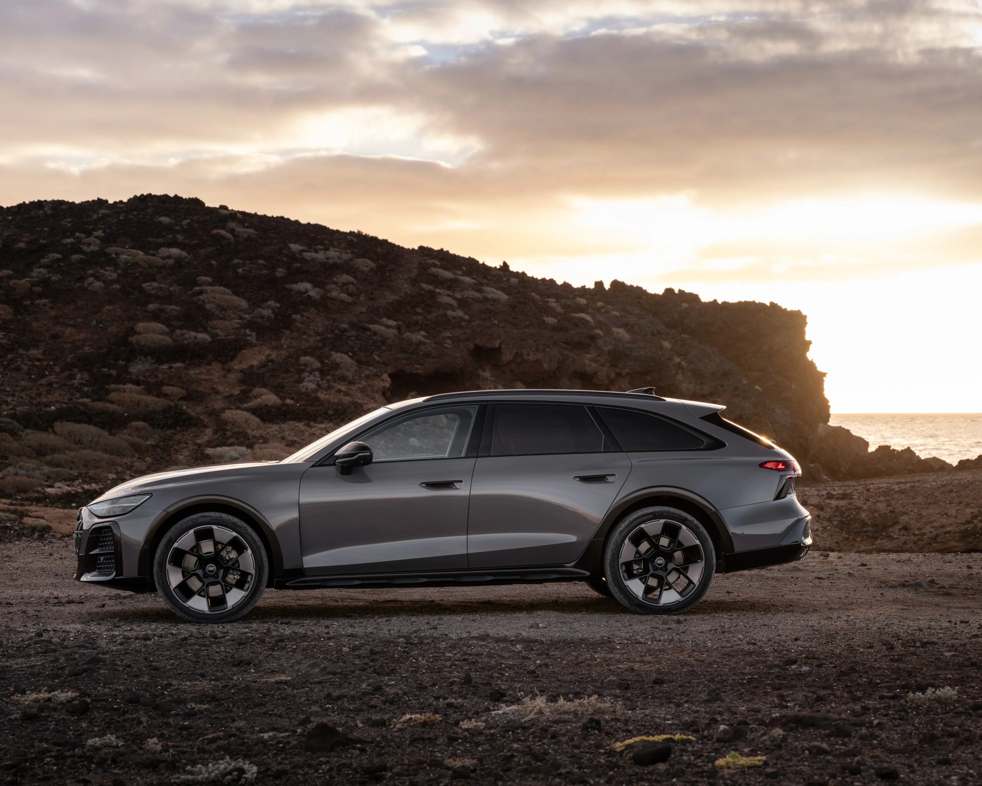 Gray Audi station wagon parked on rocky terrain with a mountainous background at sunset.