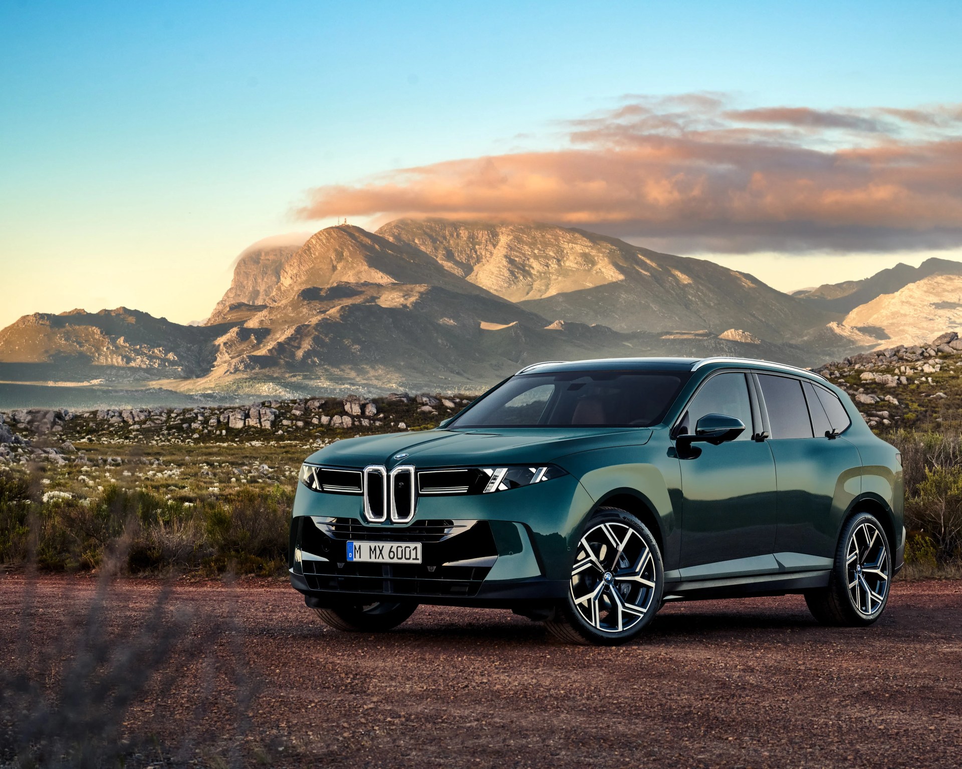 Dark green BMW SUV parked on a dirt road with rocky mountains and a cloudy sky in the background.