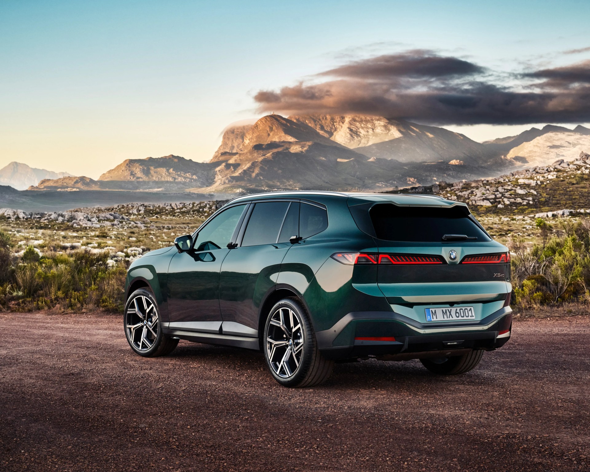 Dark green BMW X5 xDrive parked on a dirt road with rocky mountains and cloudy sky in the background.