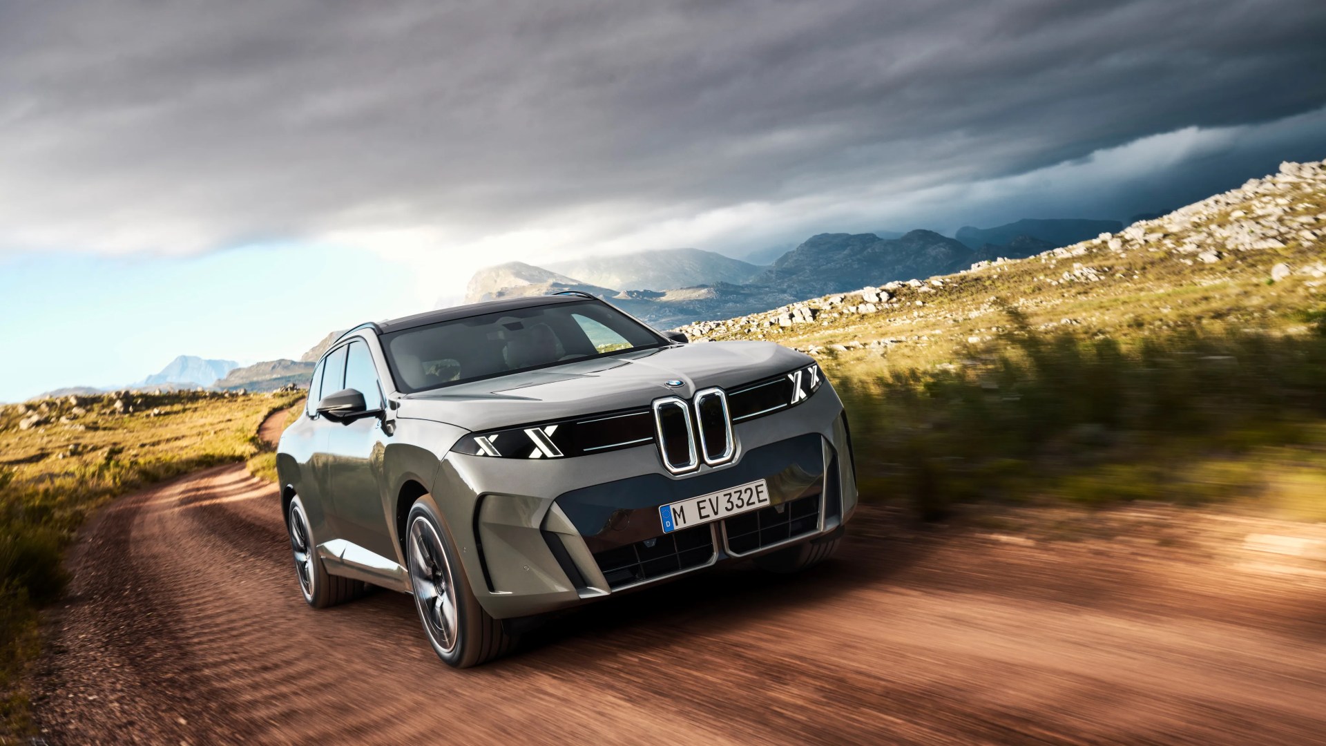 Gray BMW SUV driving on a dirt road with mountainous landscape and cloudy sky in the background.