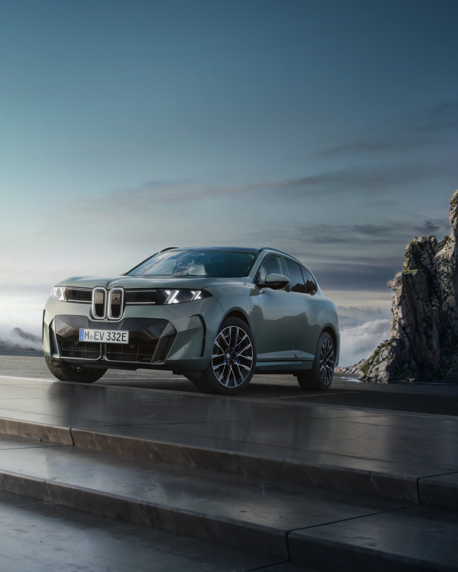 Gray BMW SUV with illuminated headlights parked on a concrete platform near rocky cliffs under a cloudy sky.