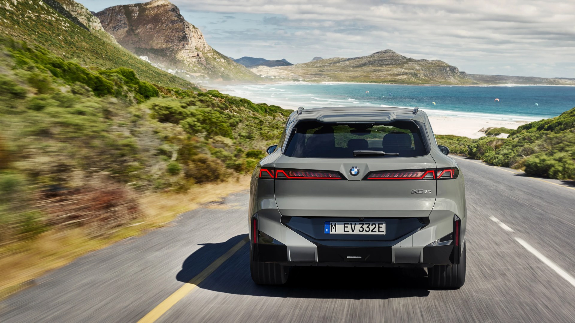 Rear view of a gray BMW iX5 electric SUV driving on a coastal road with mountains and ocean in the background.