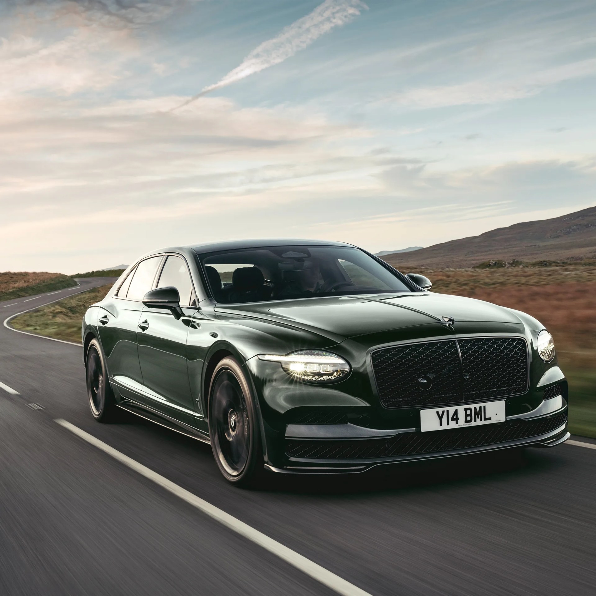 Dark green luxury sedan driving on a winding road with hills and cloudy sky in the background.