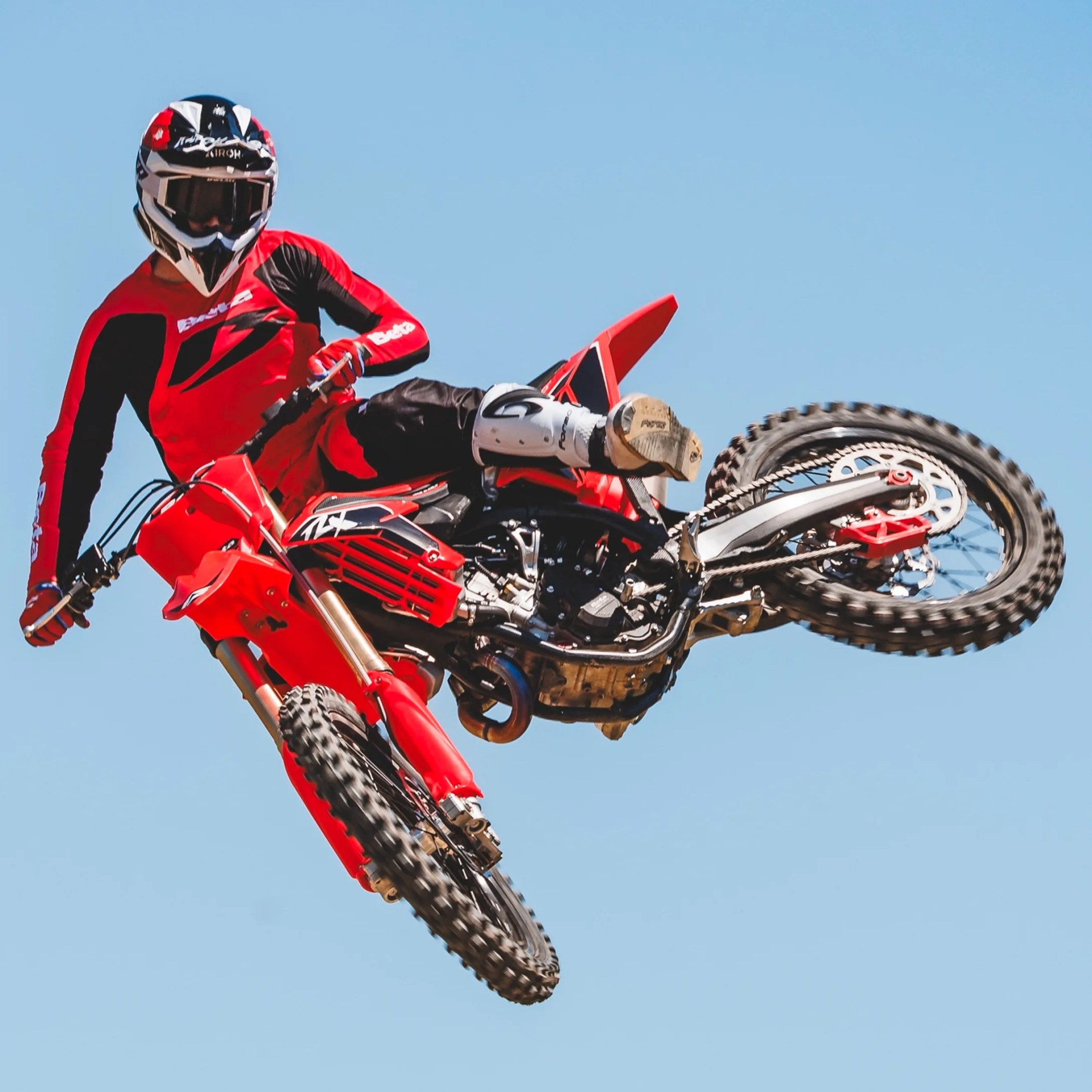 Motocross rider in red and black gear performing a jump on a red dirt bike against a clear blue sky.