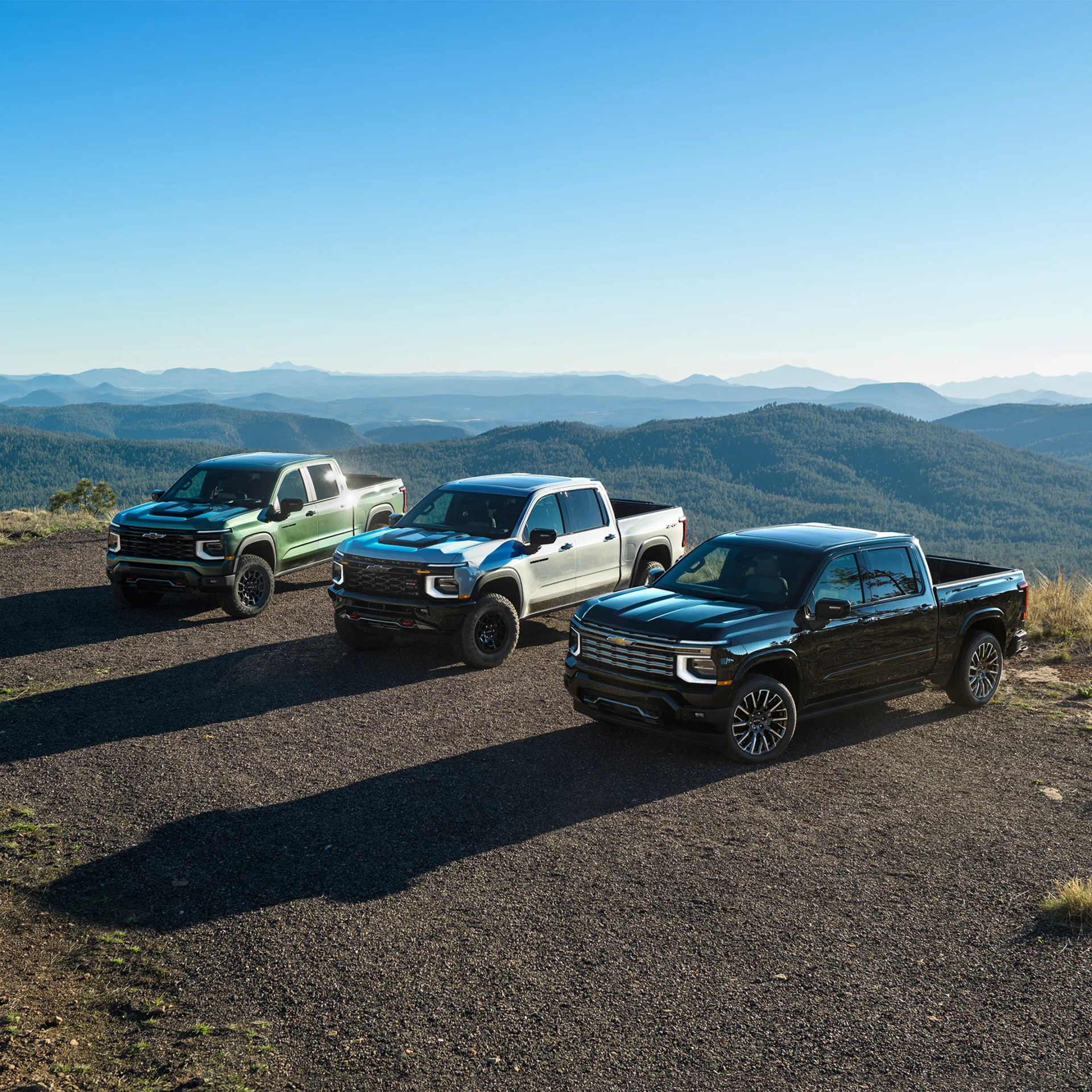Three Chevrolet Silverado pickup trucks in green, silver, and black parked on a gravel area with forested mountains in the background.