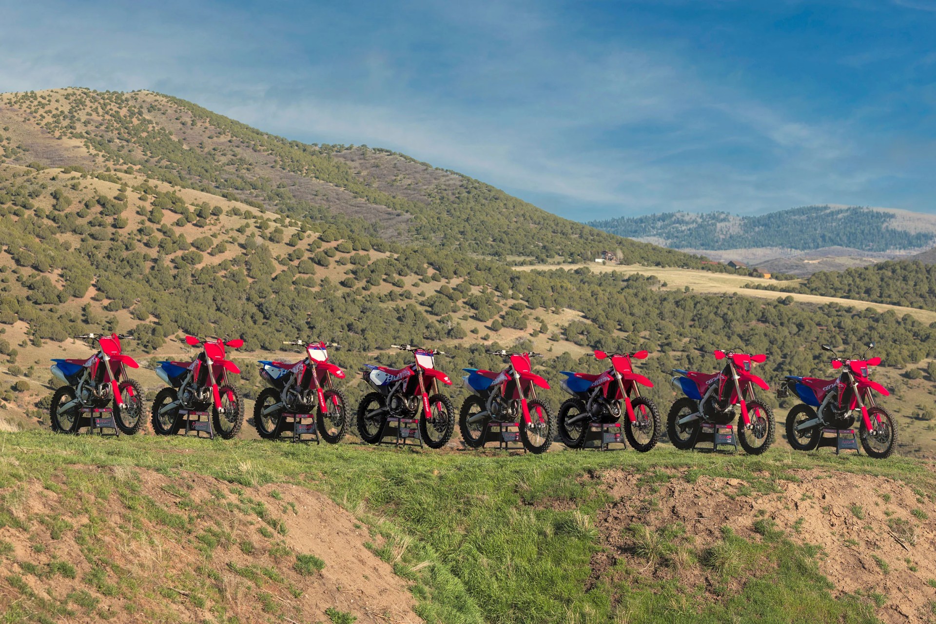 Seven red and blue Honda dirt bikes lined up on grass with forested hills and a blue sky in the background.