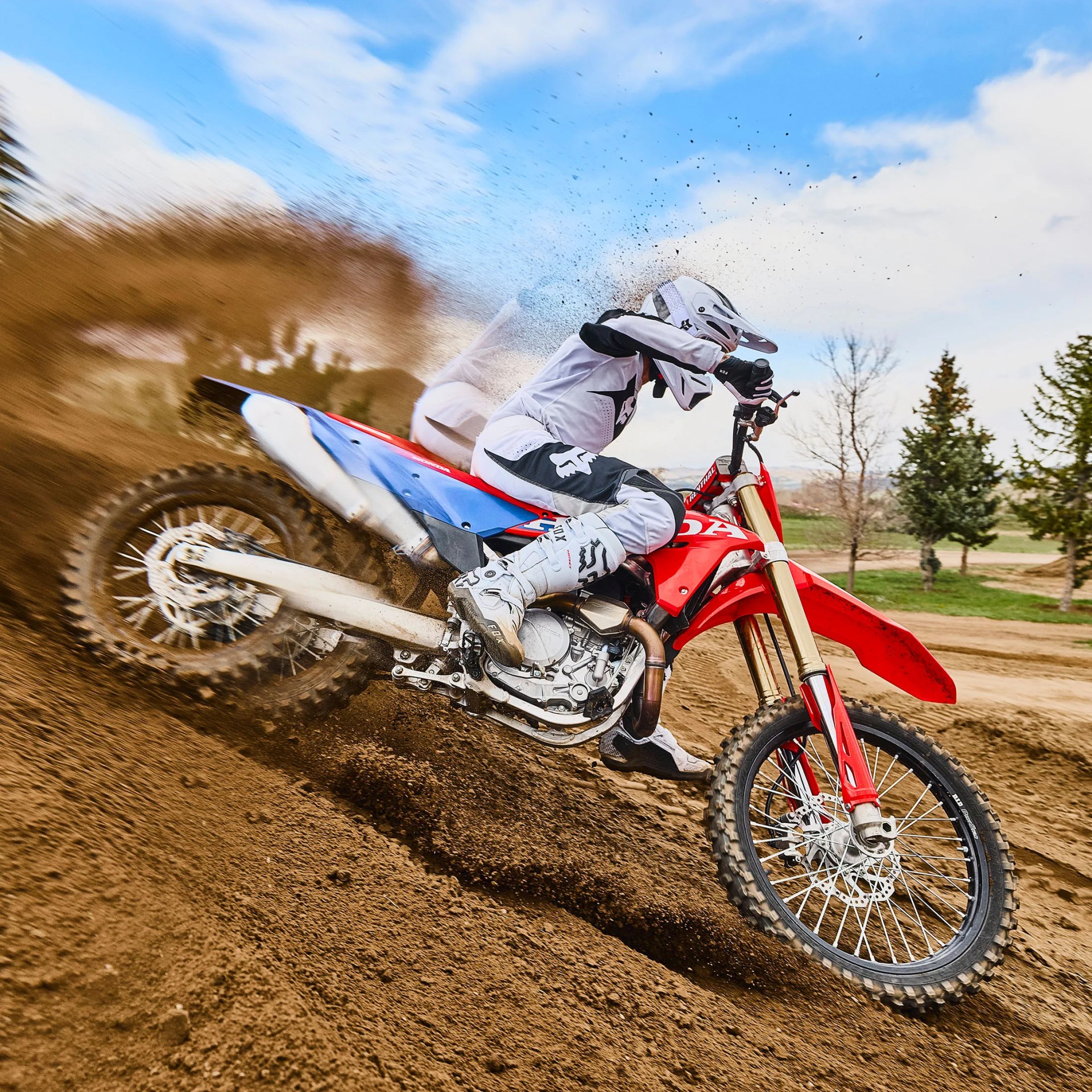 Motocross rider in white gear kicking up dirt on a red Honda dirt bike on a dirt track.
