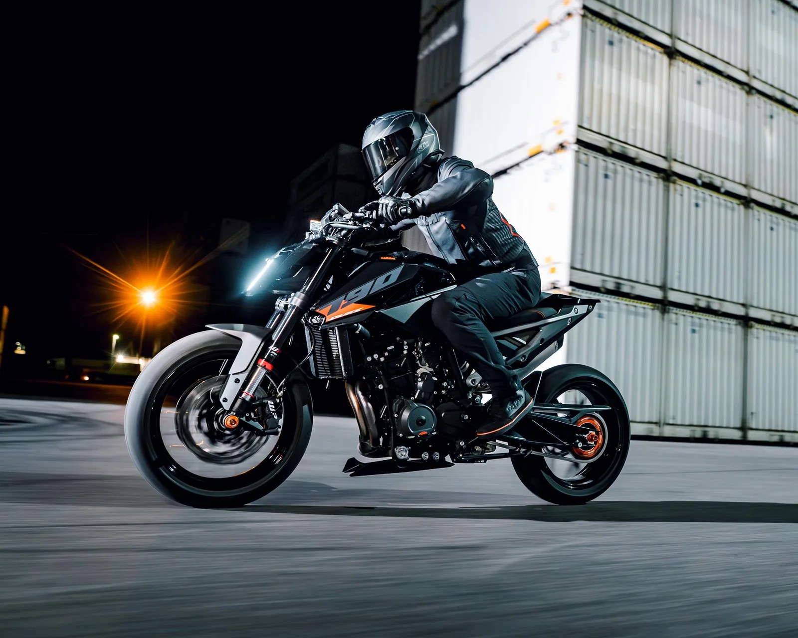 Person wearing black riding gear and helmet riding a black KTM 790 motorcycle at night near stacked shipping containers.