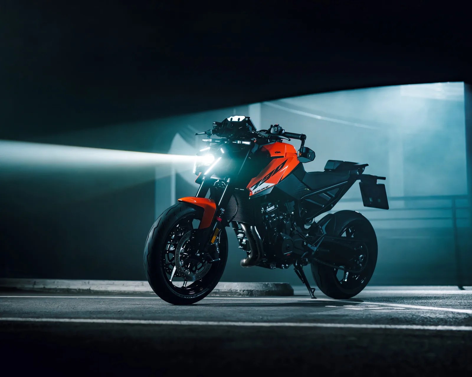 Orange and black KTM motorcycle with headlight on in a dimly lit parking garage.