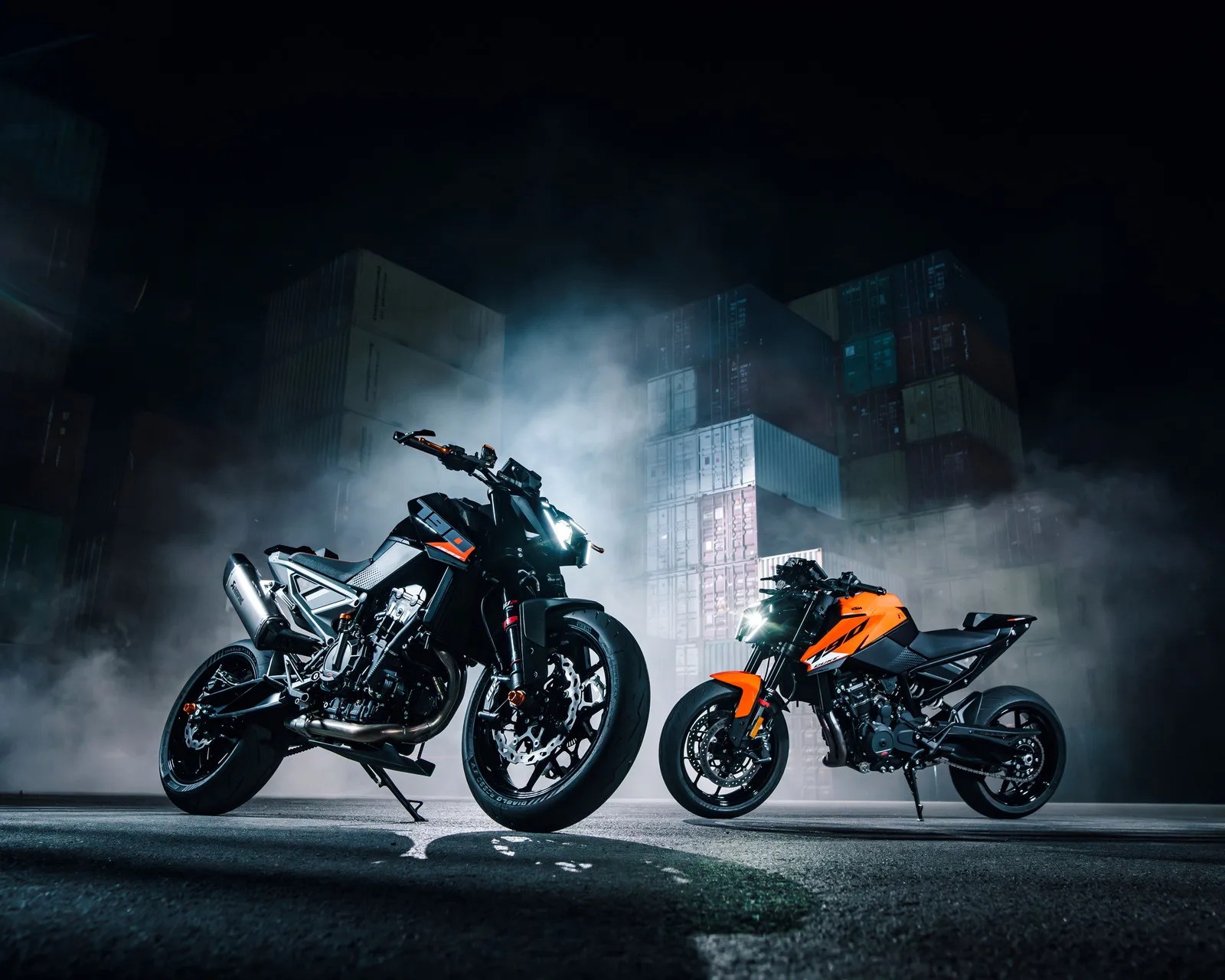 Two sport motorcycles, one black and one orange, parked on asphalt with fog and stacked shipping containers in the background at night.