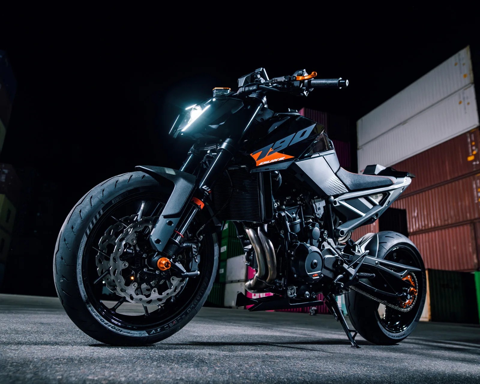 Black KTM Duke 790 motorcycle with orange accents parked on concrete near stacked shipping containers at night.