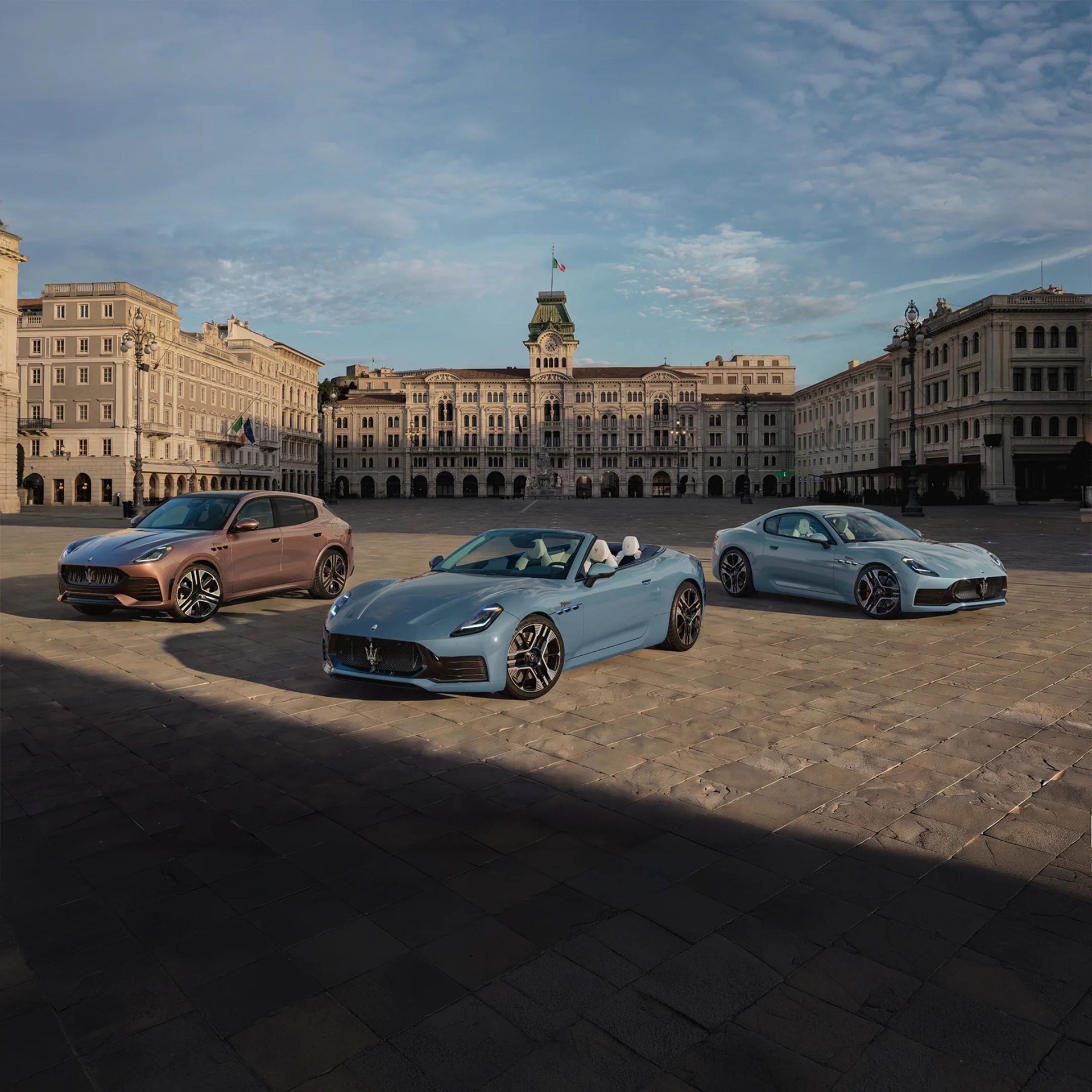Three Maserati cars in blue and bronze colors parked on a large stone plaza with historic European buildings in the background.