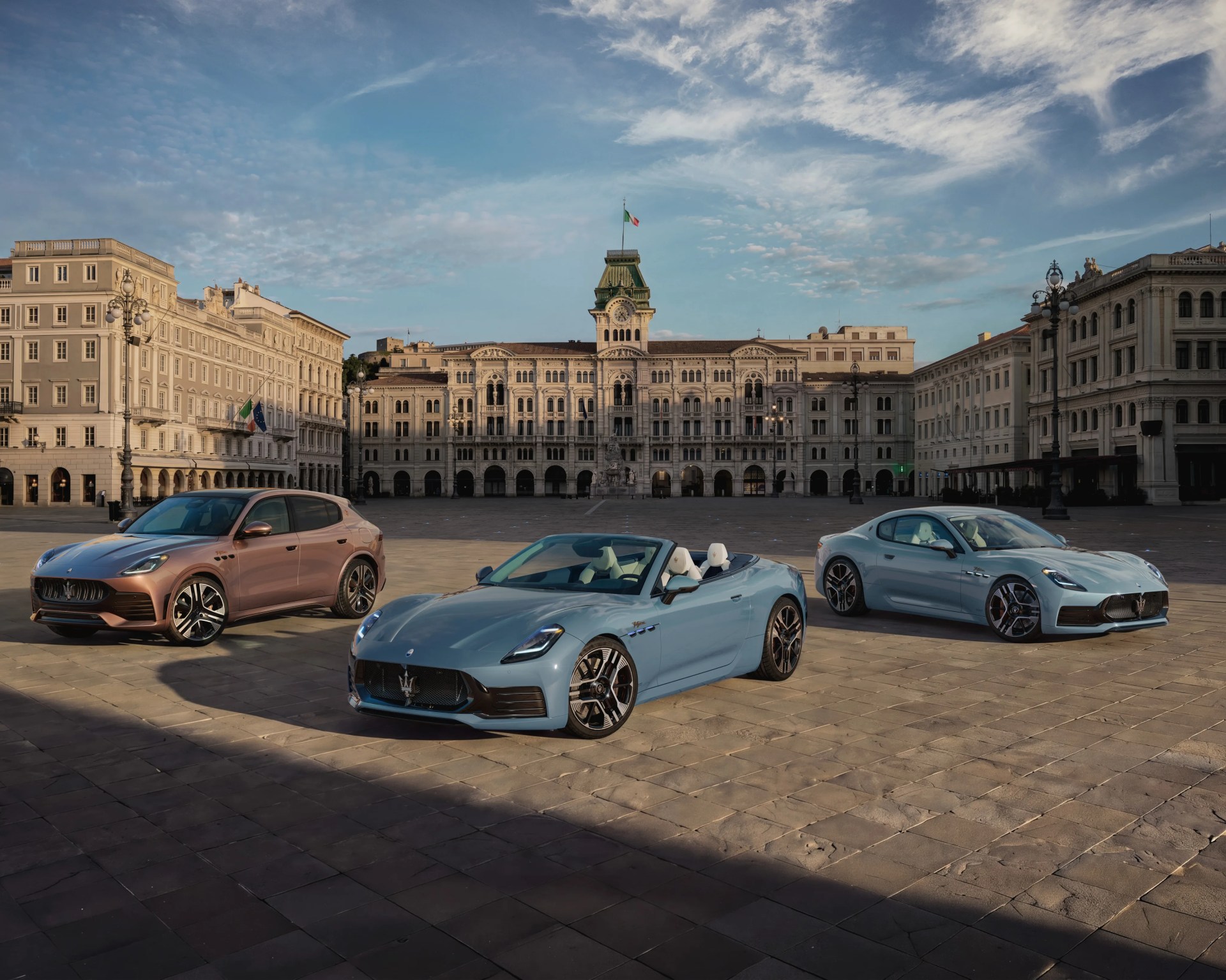 Three Maserati cars, two light blue and one bronze, parked in a large historic square with an ornate building and Italian flag in the background.