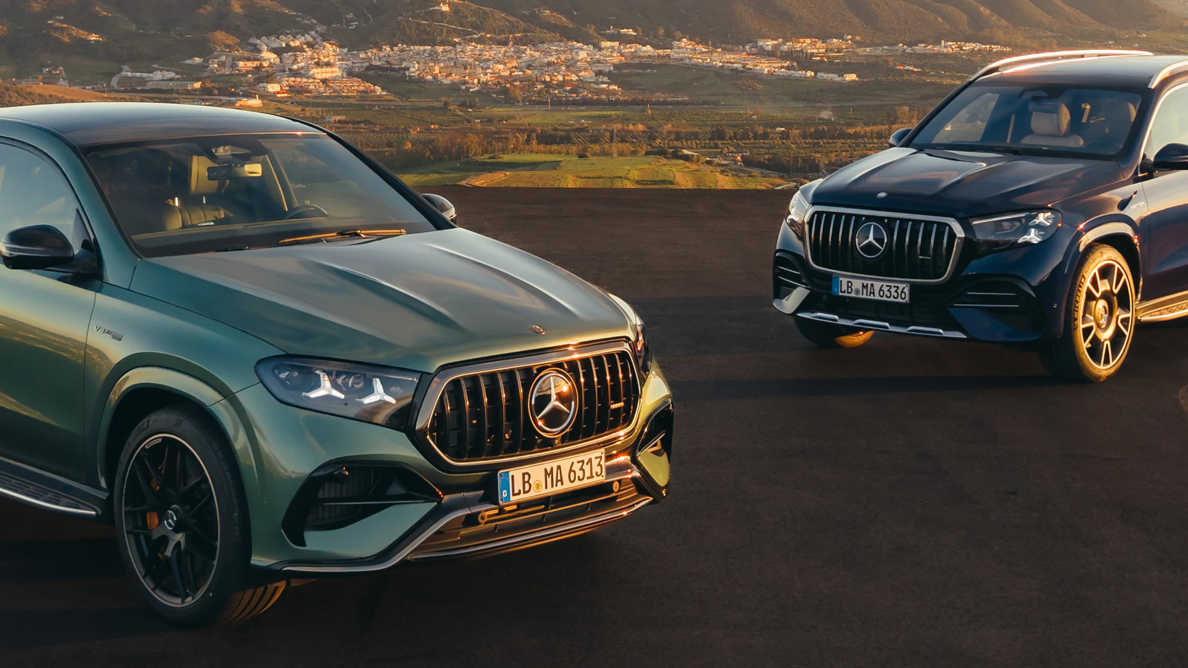 Two Mercedes-Benz SUVs, one green and one black, parked on a paved surface with a mountainous town in the background.