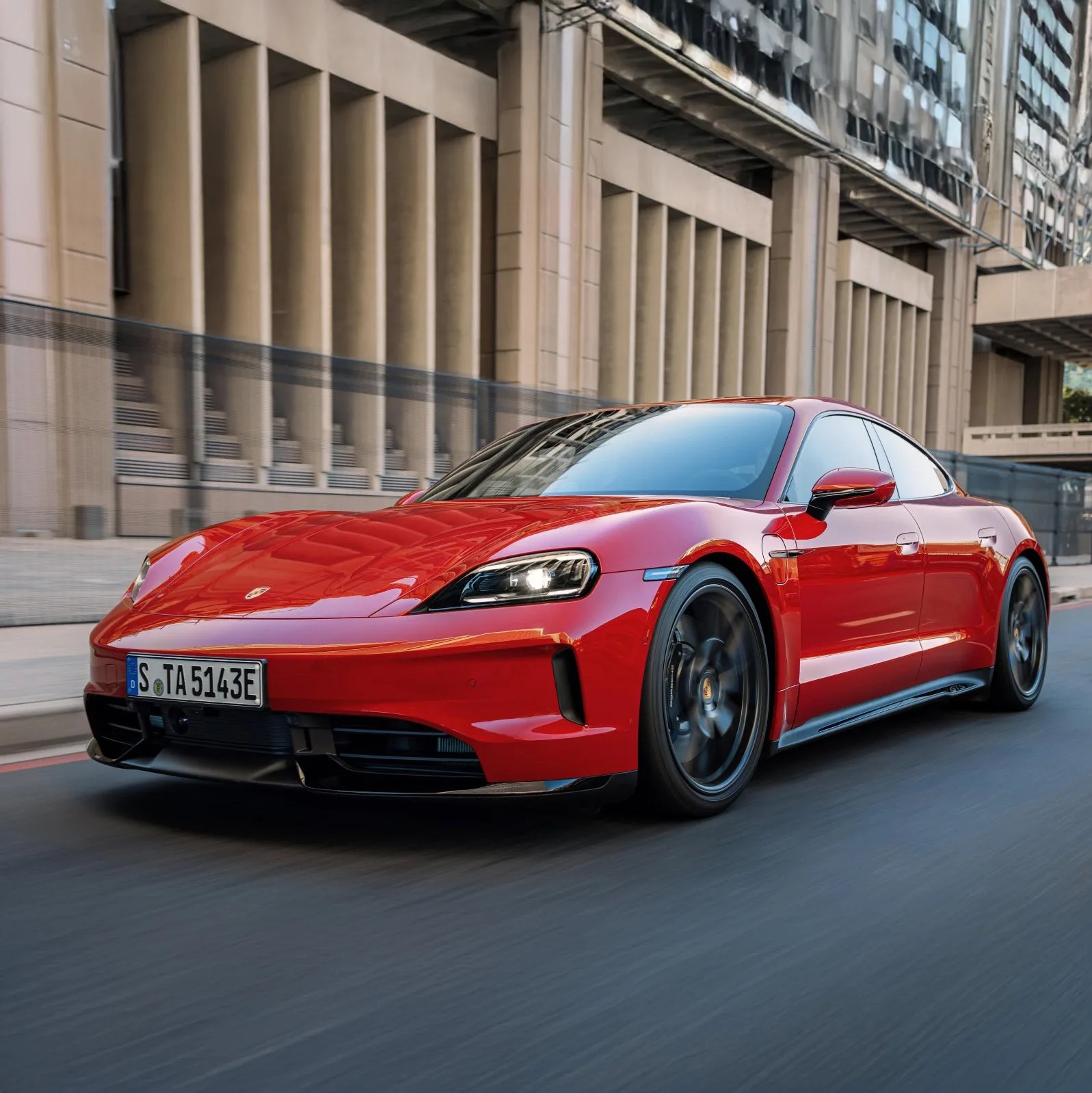 Red Porsche Taycan GTS electric sports car driving on a city street with modern buildings in the background.