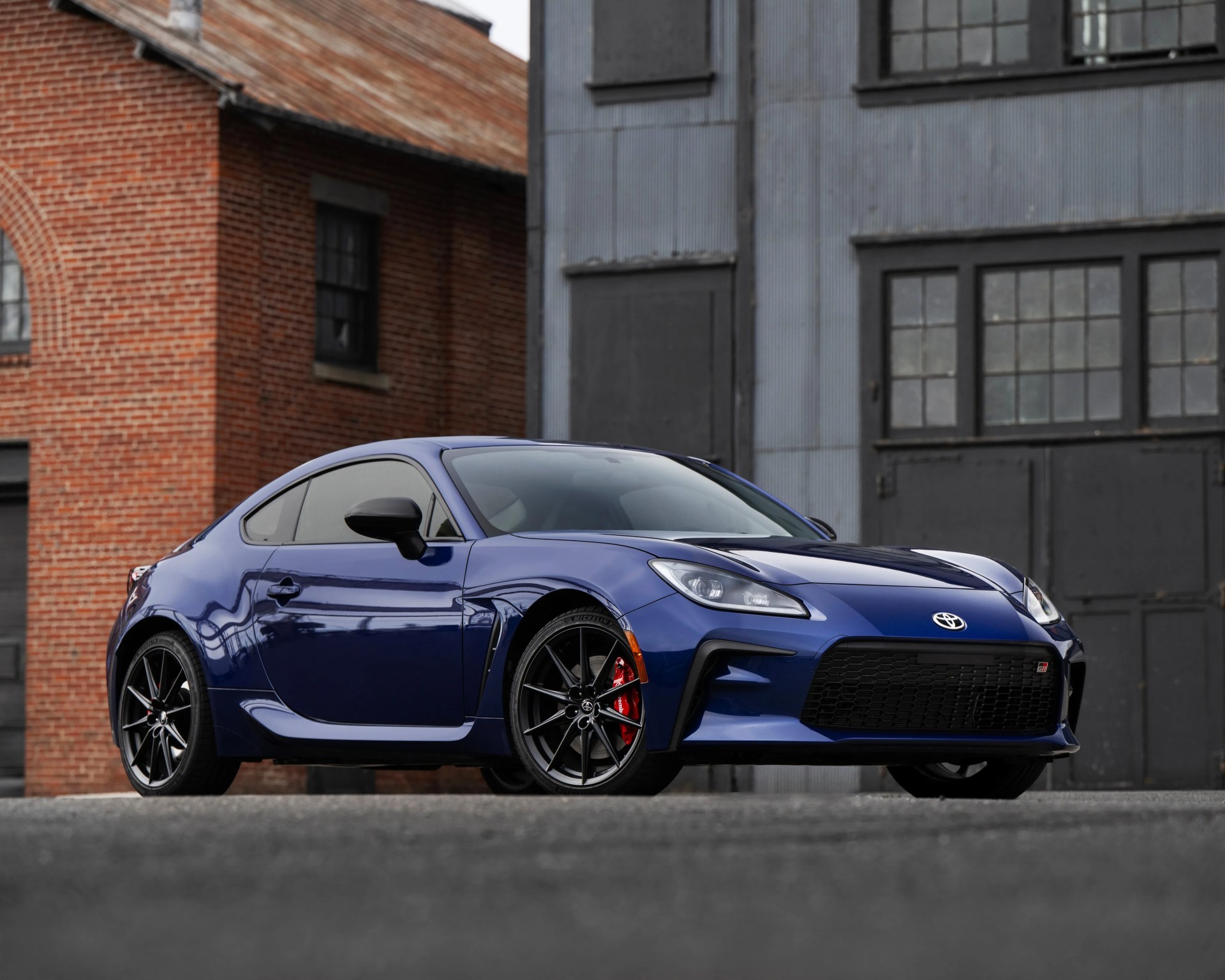Blue Toyota GR sports car with black wheels and red brake calipers parked in front of brick and metal buildings.