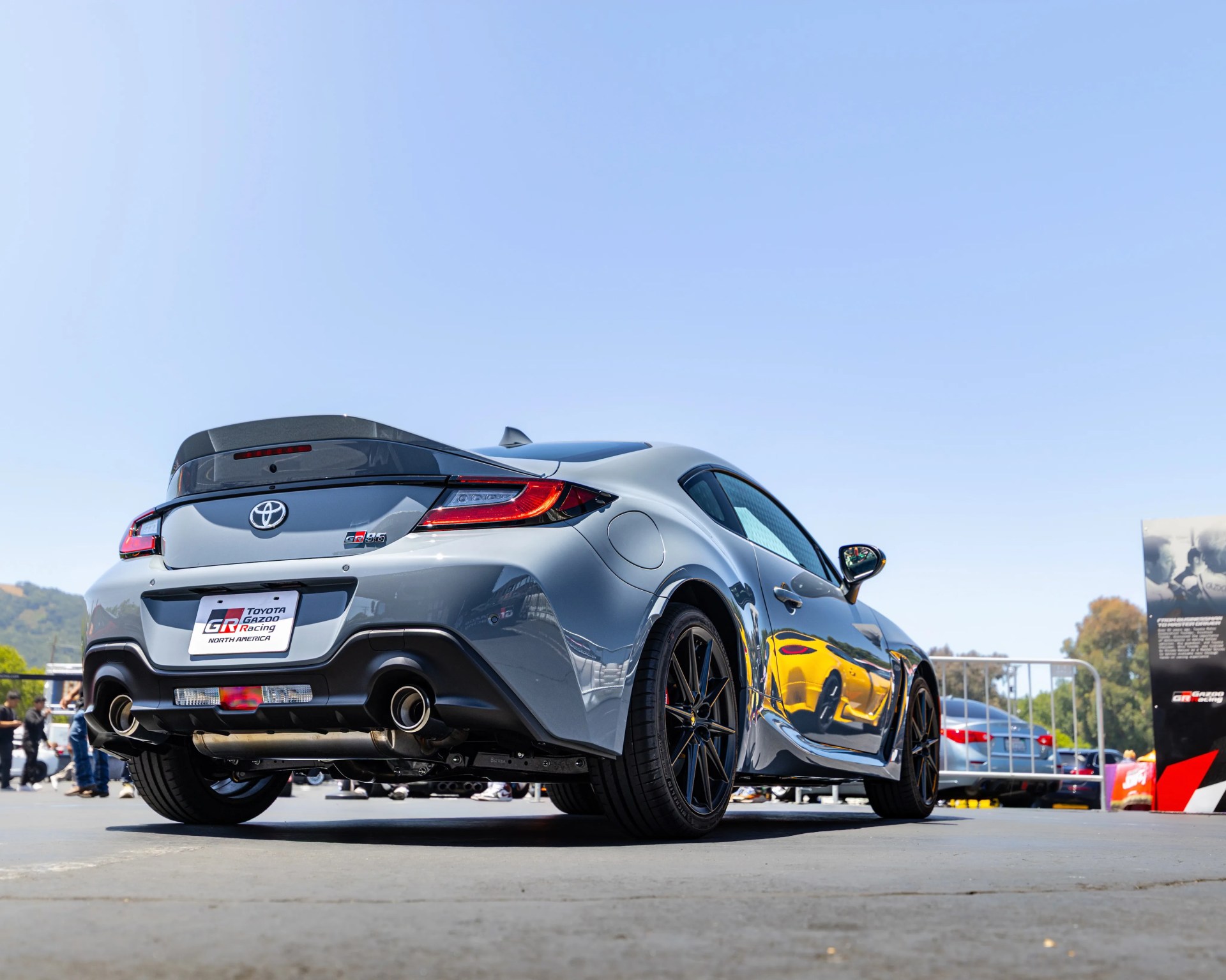 Gray Toyota GR86 sports car rear with black spoiler and dual exhausts under clear blue sky.