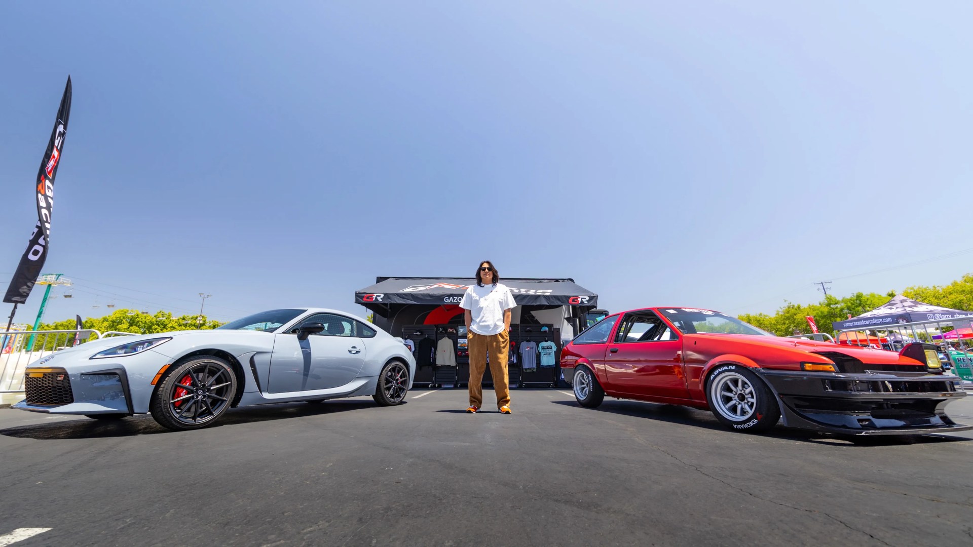 Person standing between a silver sports car and a red vintage race car in front of a Gazoo Racing tent.