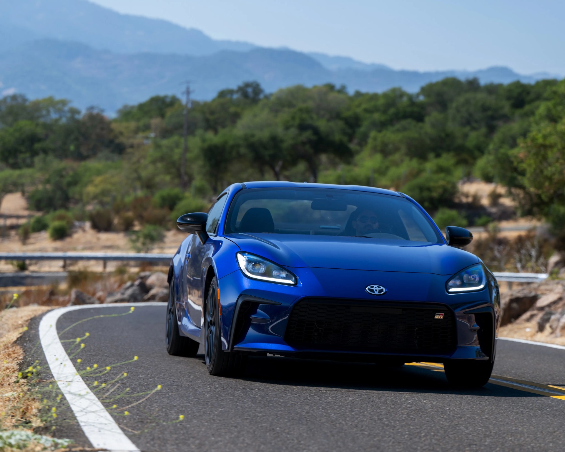 Blue Toyota GR sports car driving on a winding road with trees and mountains in the background.