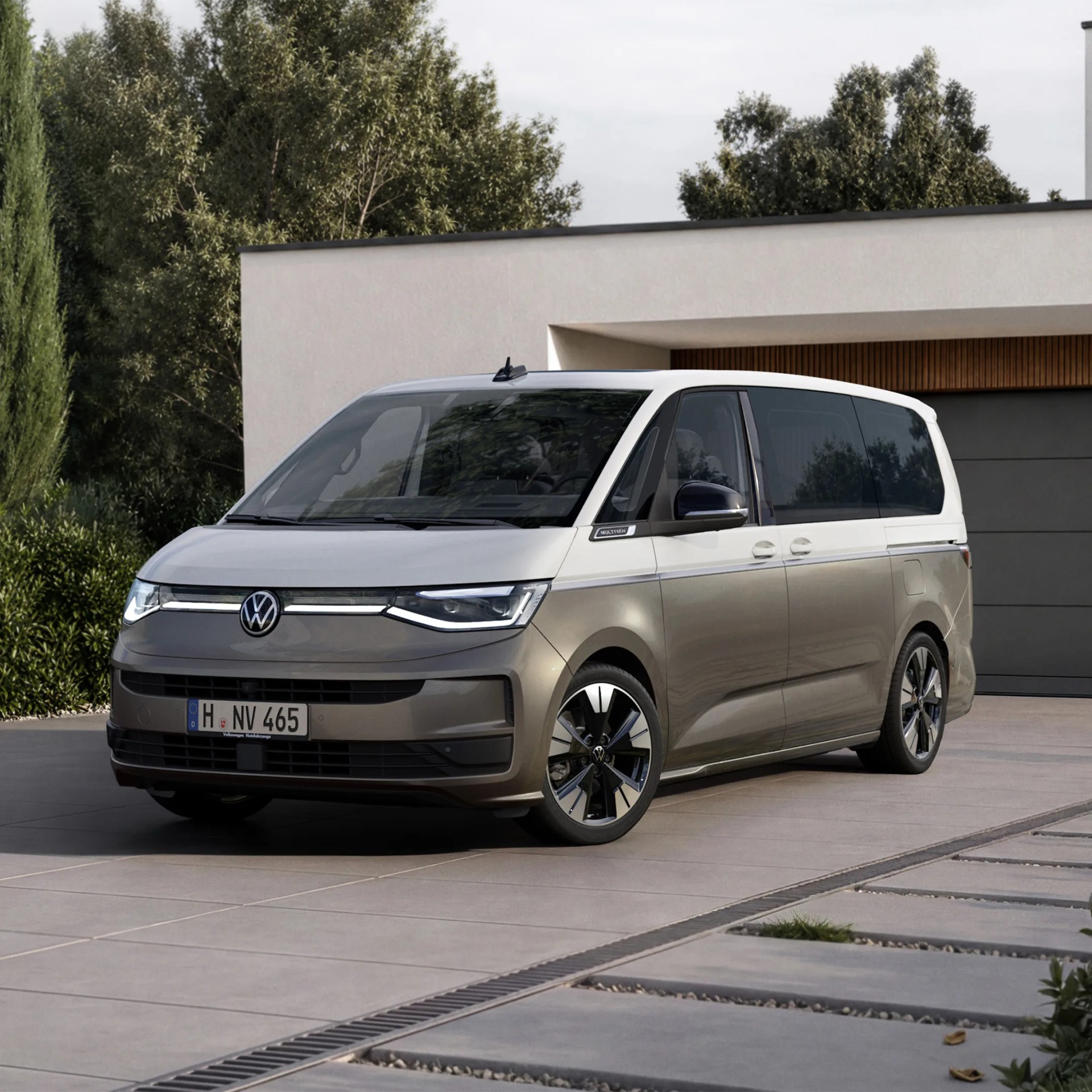 Two-tone beige and white Volkswagen Multivan parked on a tiled driveway in front of a modern house.