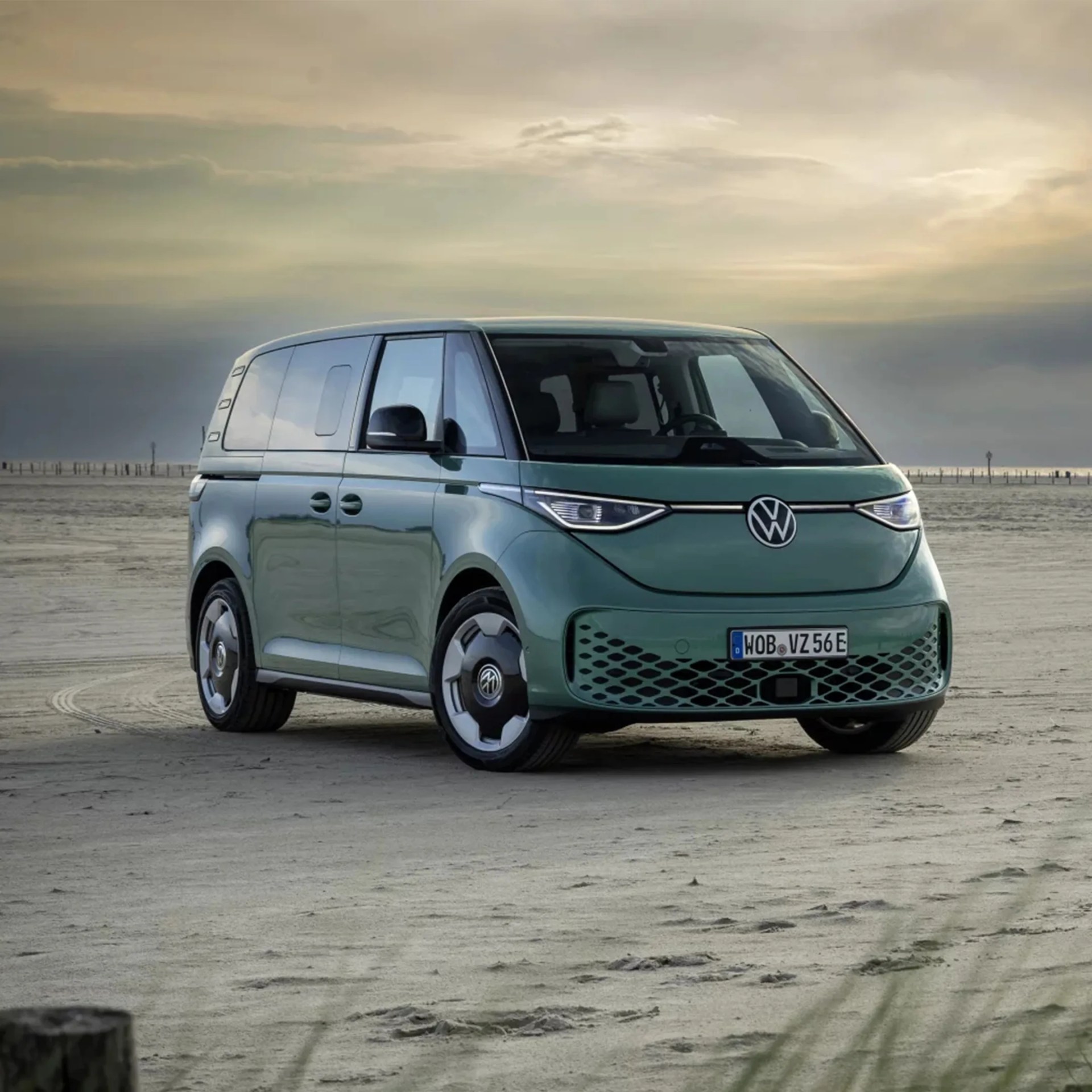Green Volkswagen electric van parked on a sandy beach under a cloudy sky.