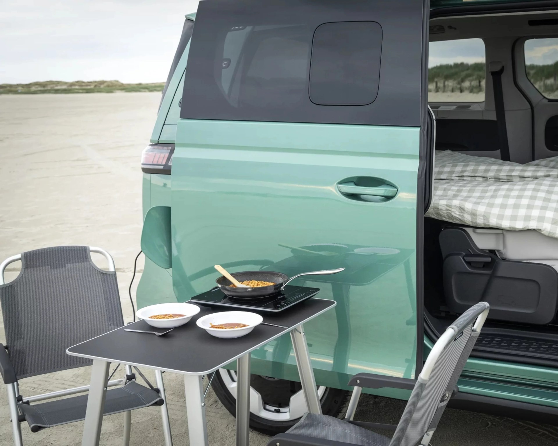 Teal camper van with sliding door open showing a bed inside, a black folding table with two bowls and a frying pan on a portable stove, and two gray folding chairs on a sandy beach.