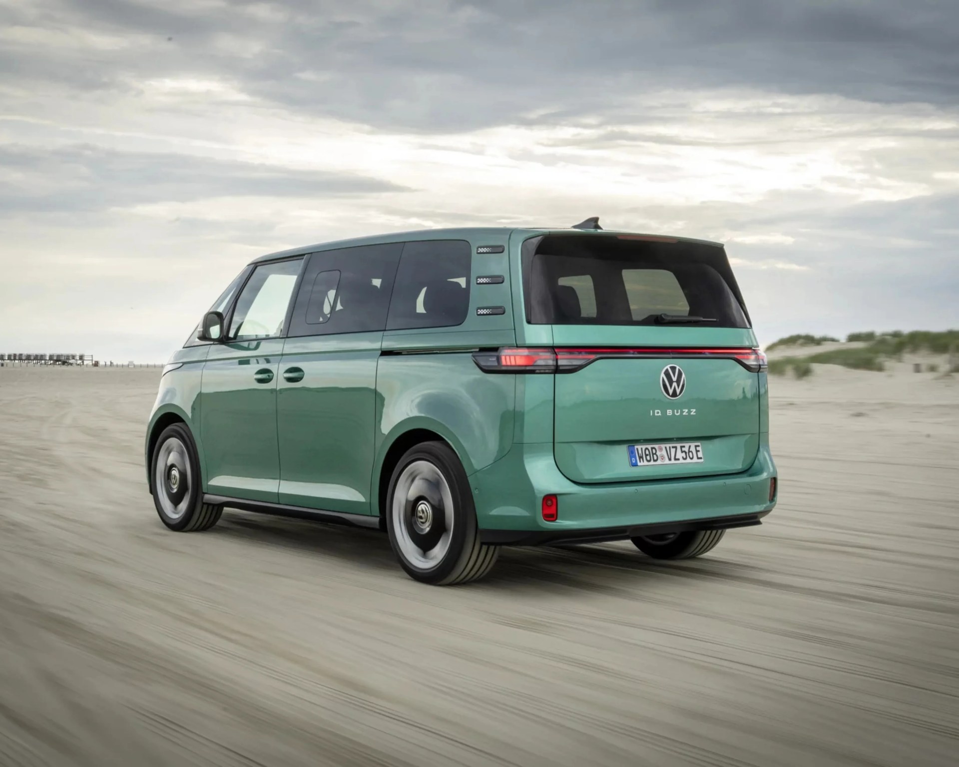 Green Volkswagen ID. Buzz electric van driving on a sandy beach under a cloudy sky.