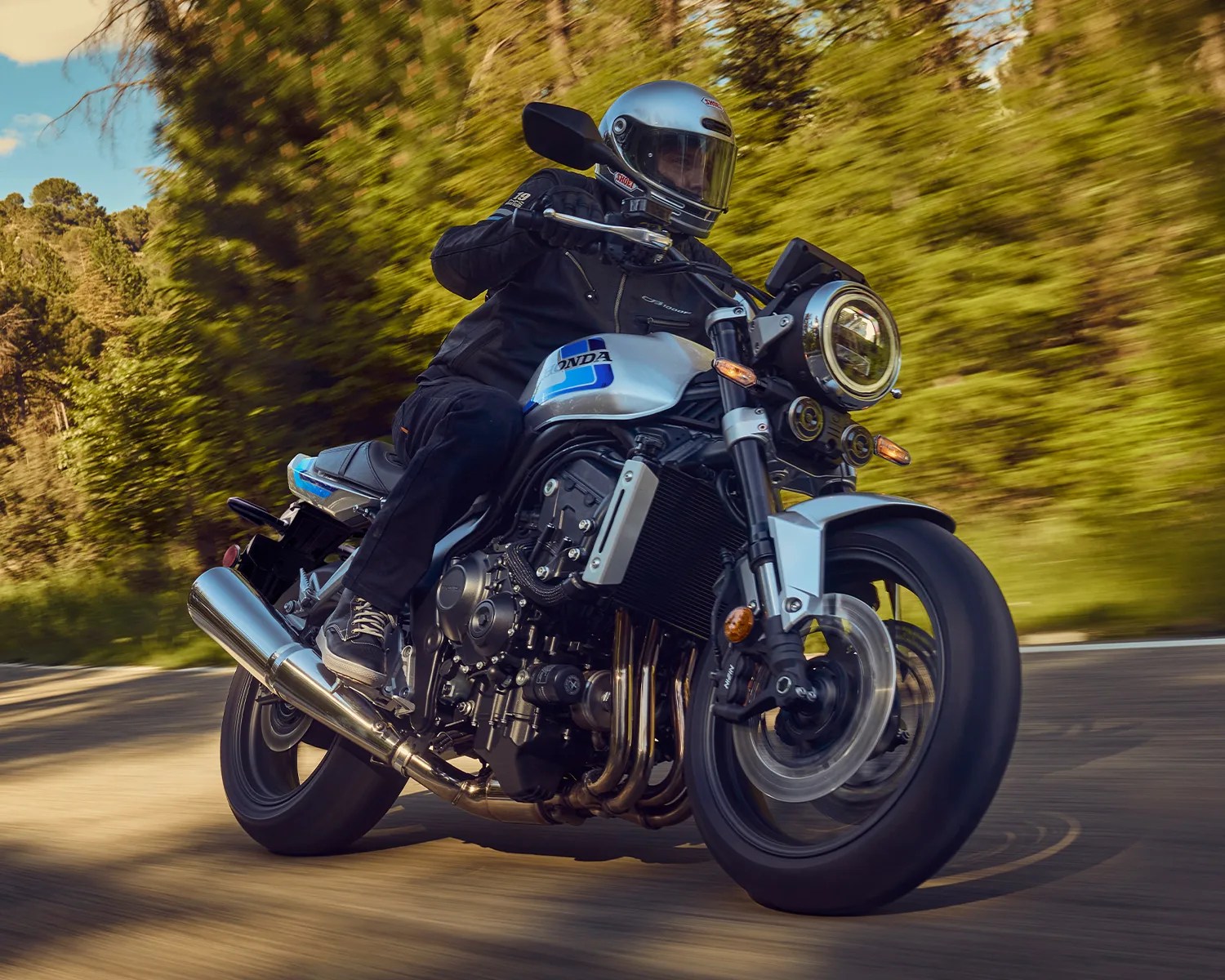 Silver Honda motorcycle with blue accents ridden on a curving road surrounded by green trees.