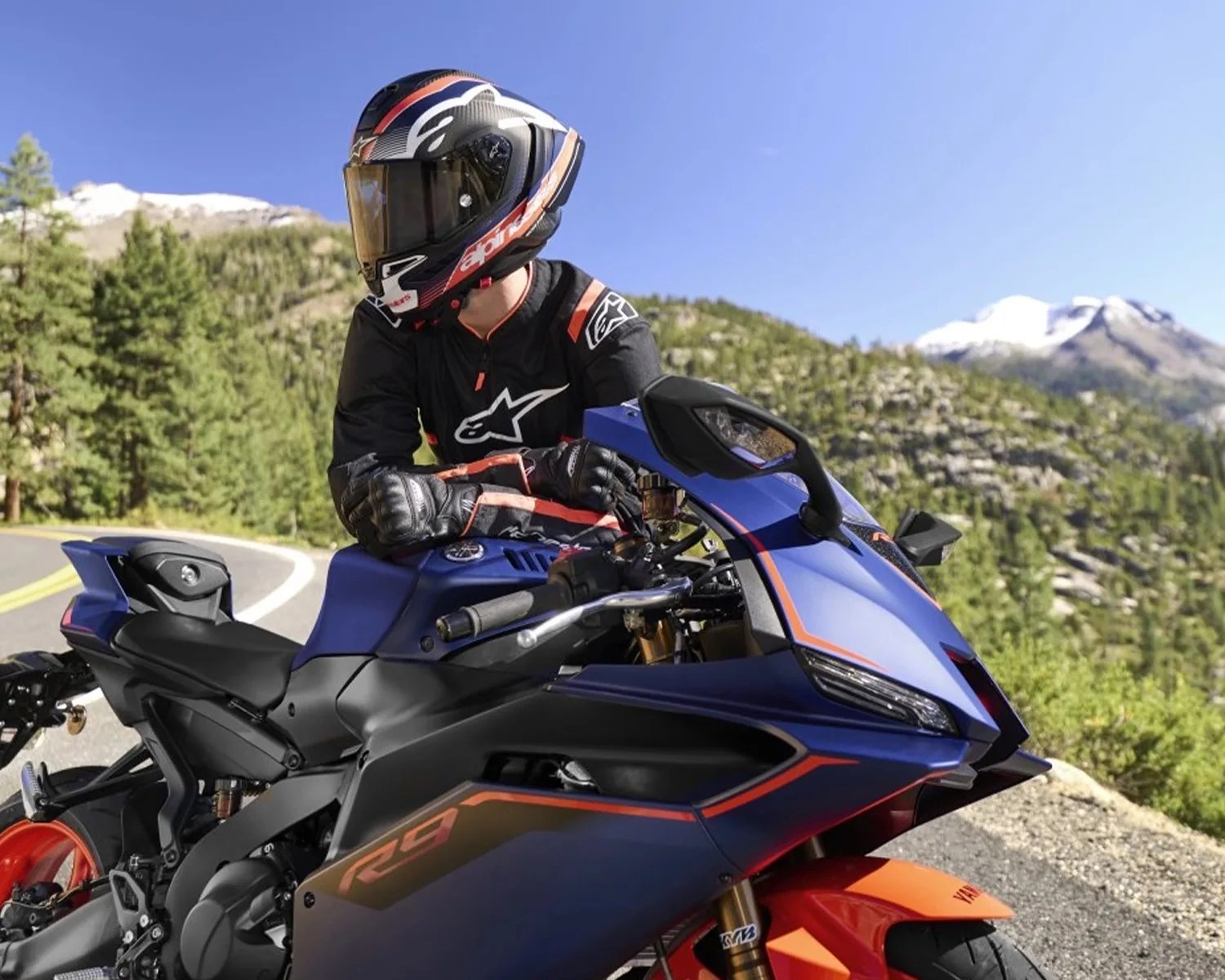 Person wearing a black and orange motorcycle helmet and gear sitting on a blue and orange Yamaha R9 sportbike on a mountain road.