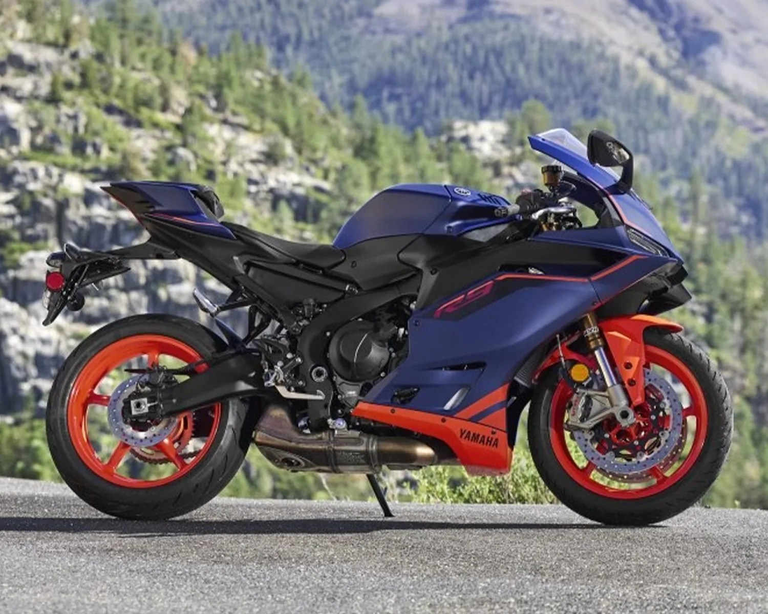Blue and orange Yamaha sport motorcycle with orange wheels parked on a road with a mountainous background.