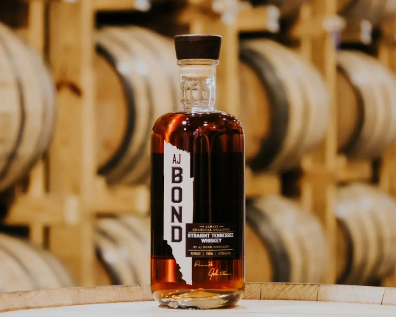 Bottle of AJ Bond Straight Tennessee Whiskey with wooden barrels in the background.