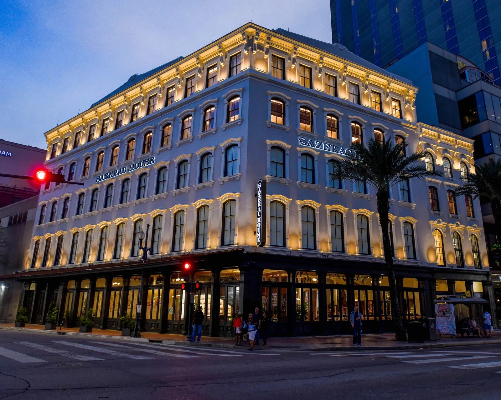 Illuminated historic Sazerac House building at dusk on a city street corner with palm trees and pedestrians.