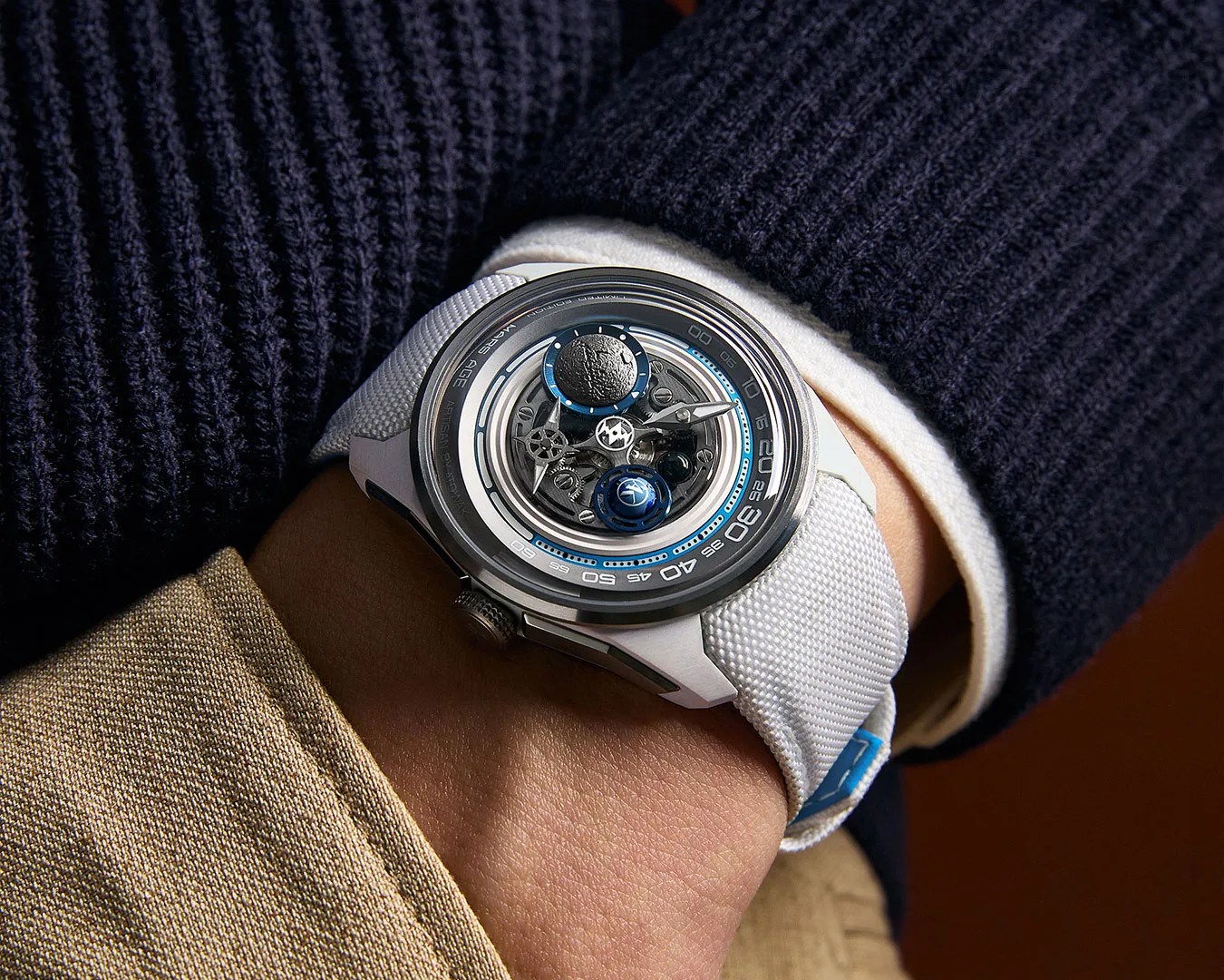 Silver wristwatch with a white textured strap and a complex mechanical dial featuring blue and black accents, worn on a wrist.