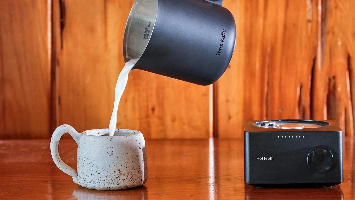 Black Terra Kaffe milk frothing pitcher pouring milk into a speckled ceramic mug next to a black Hot Froth frother on a wooden surface.