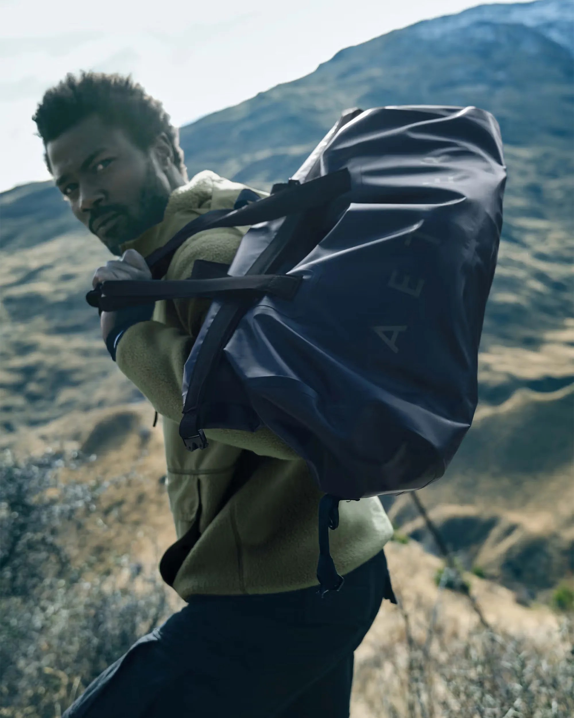 Man wearing olive-green fleece jacket carrying large black duffel bag outdoors with mountains in background.
