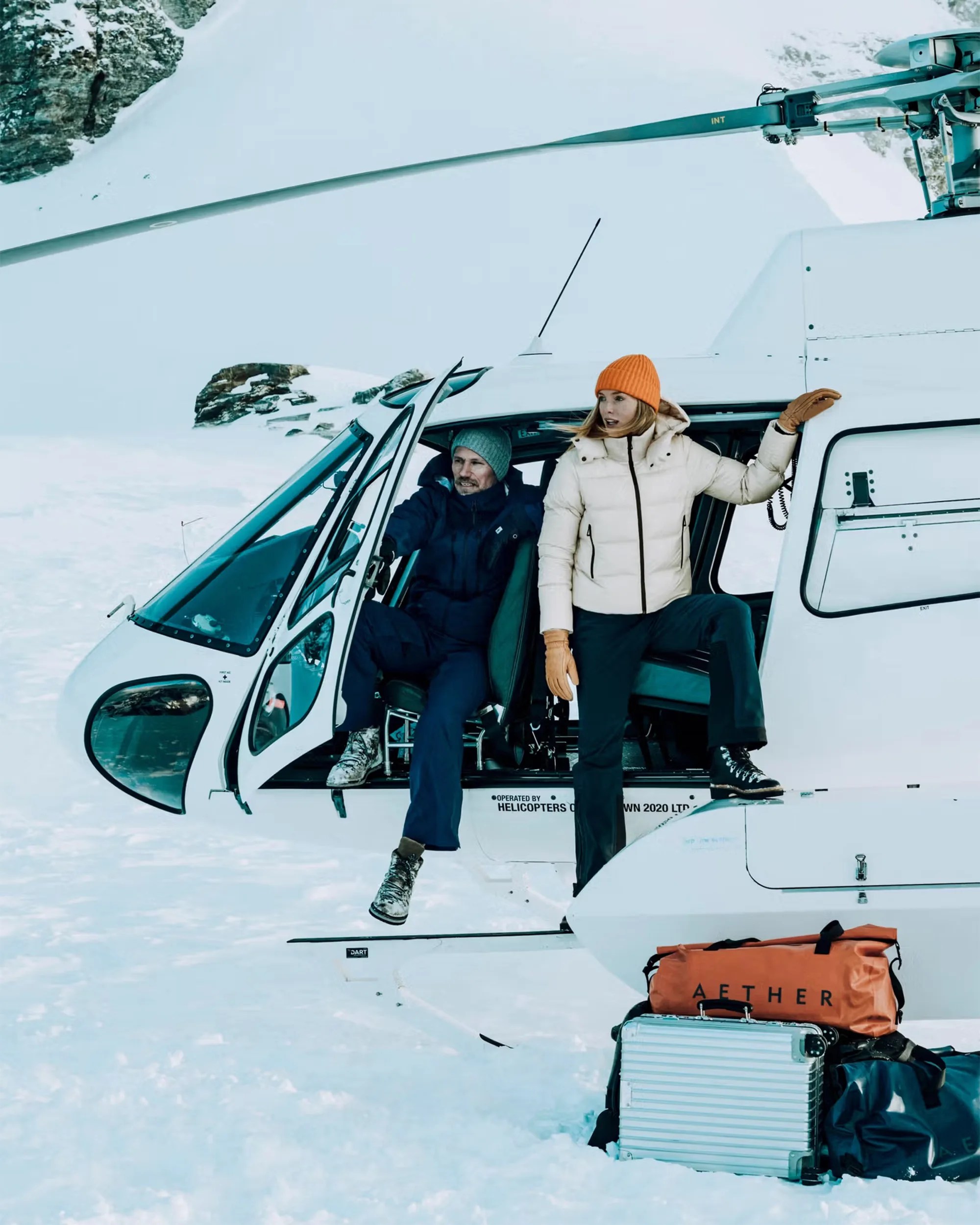 White helicopter with open door on snowy mountain, two people in winter clothing exiting, and luggage on snow.