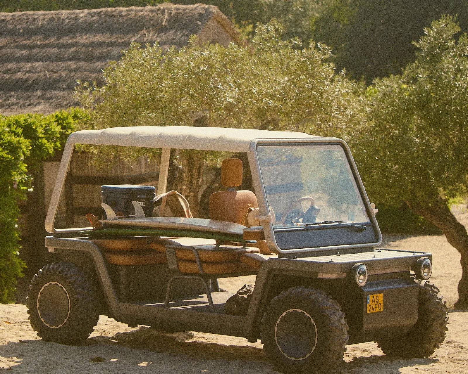 Open-top off-road vehicle with tan seats, a surfboard, and a cooler on sandy terrain near trees.