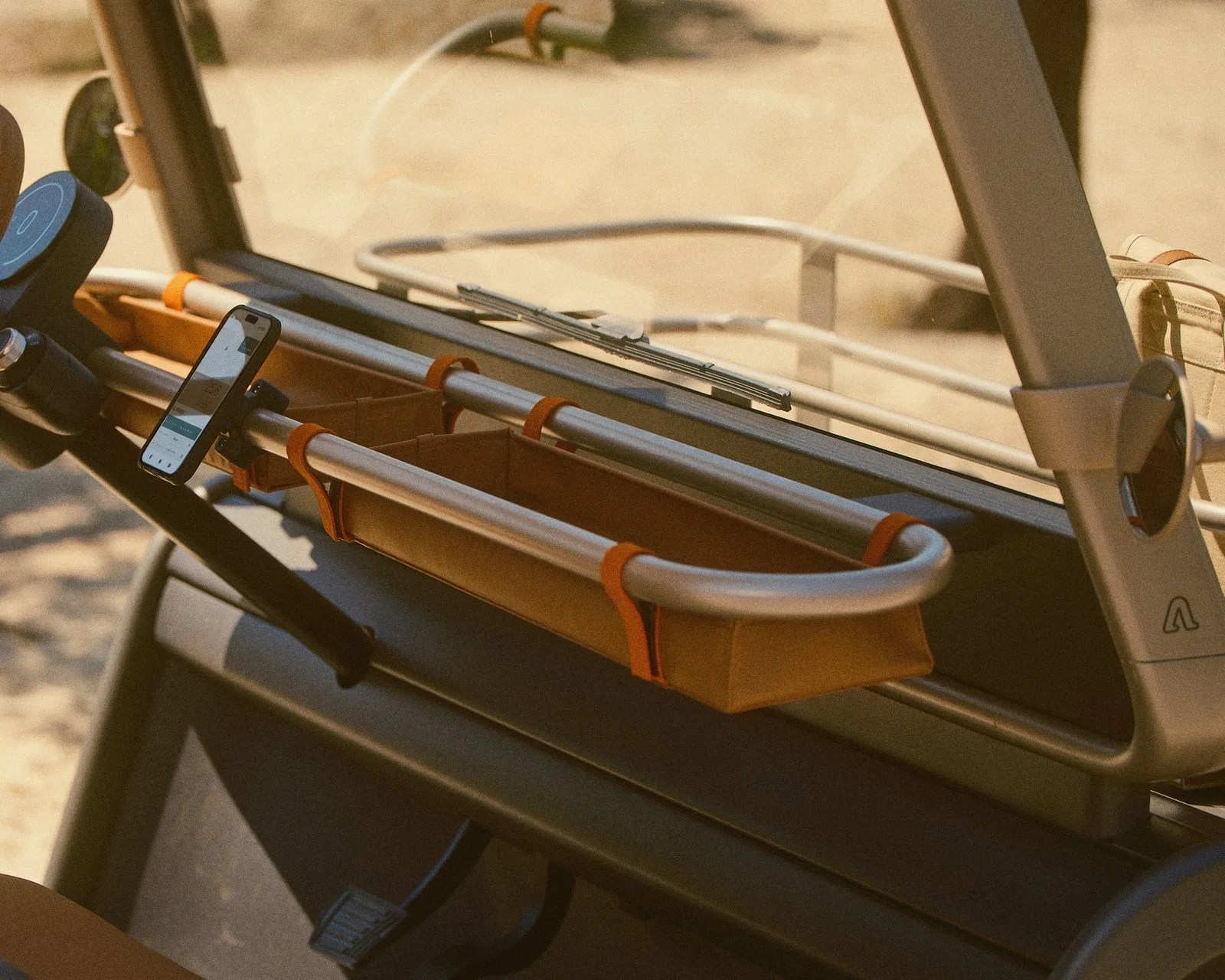 Brown fabric storage basket with orange straps attached to silver metal bars inside a vehicle dashboard.