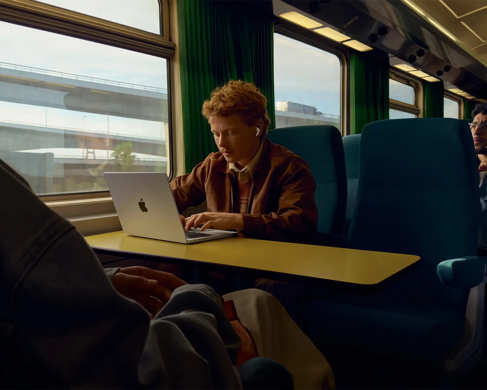 Person using a silver Apple MacBook laptop on a yellow table inside a train with green curtains and blue seats.