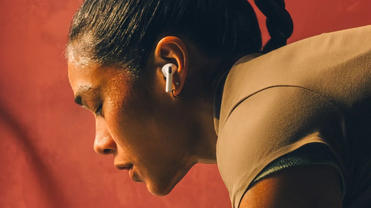Close-up of a person wearing white wireless earbuds with a braided hairstyle and a brown top against a red background.
