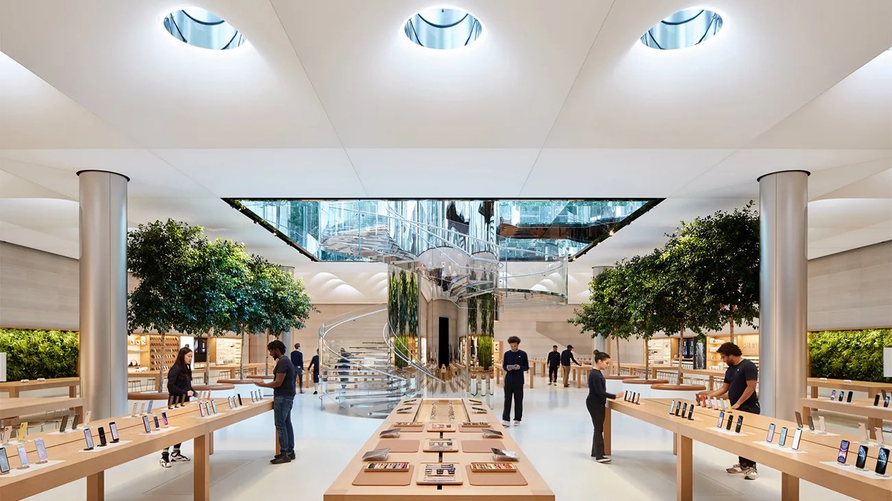 Spacious modern retail store with wooden tables displaying smartphones, large green trees, and a central glass spiral staircase.