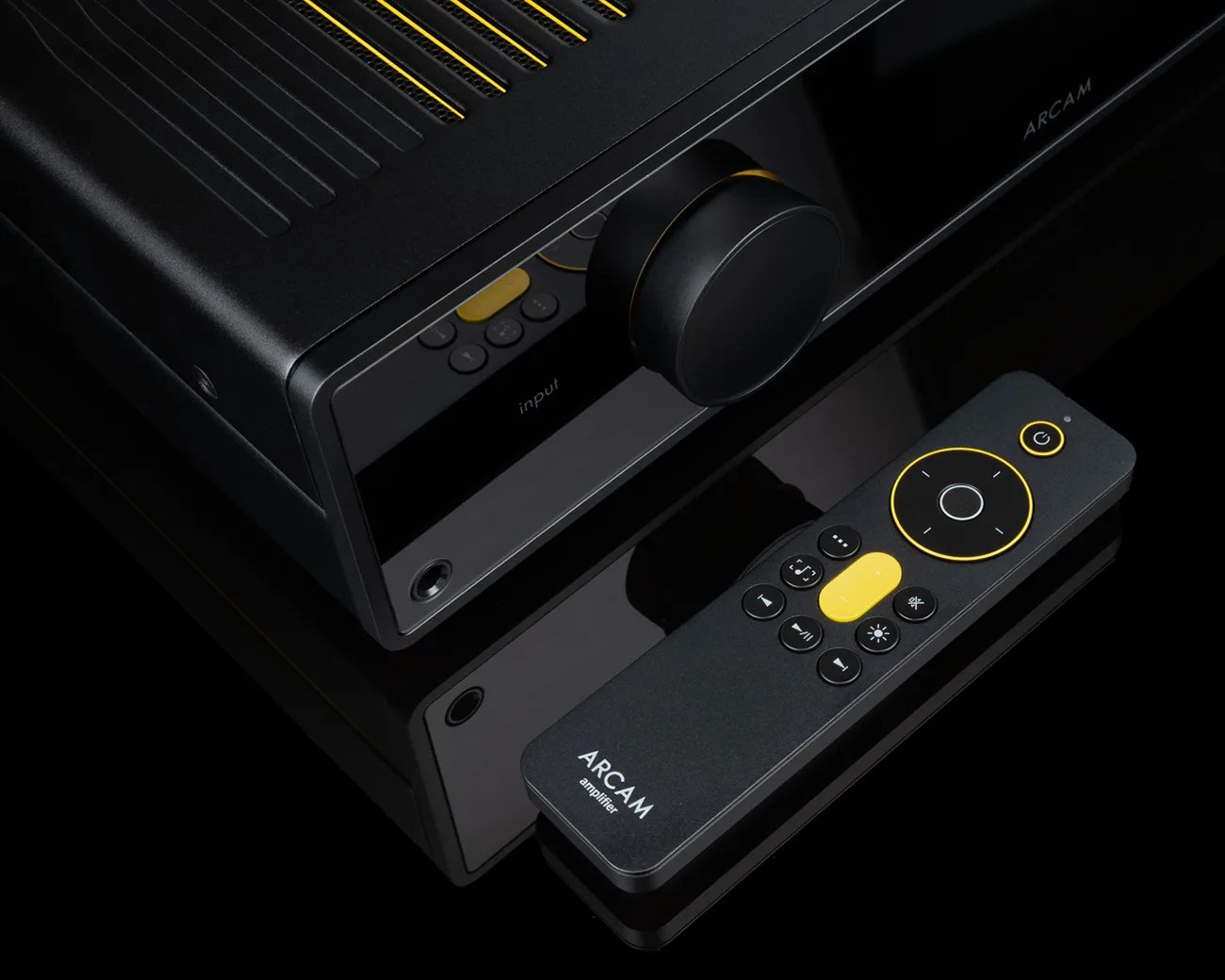 Black Arcam amplifier with large volume knob and matching black remote with yellow accents on a reflective surface.