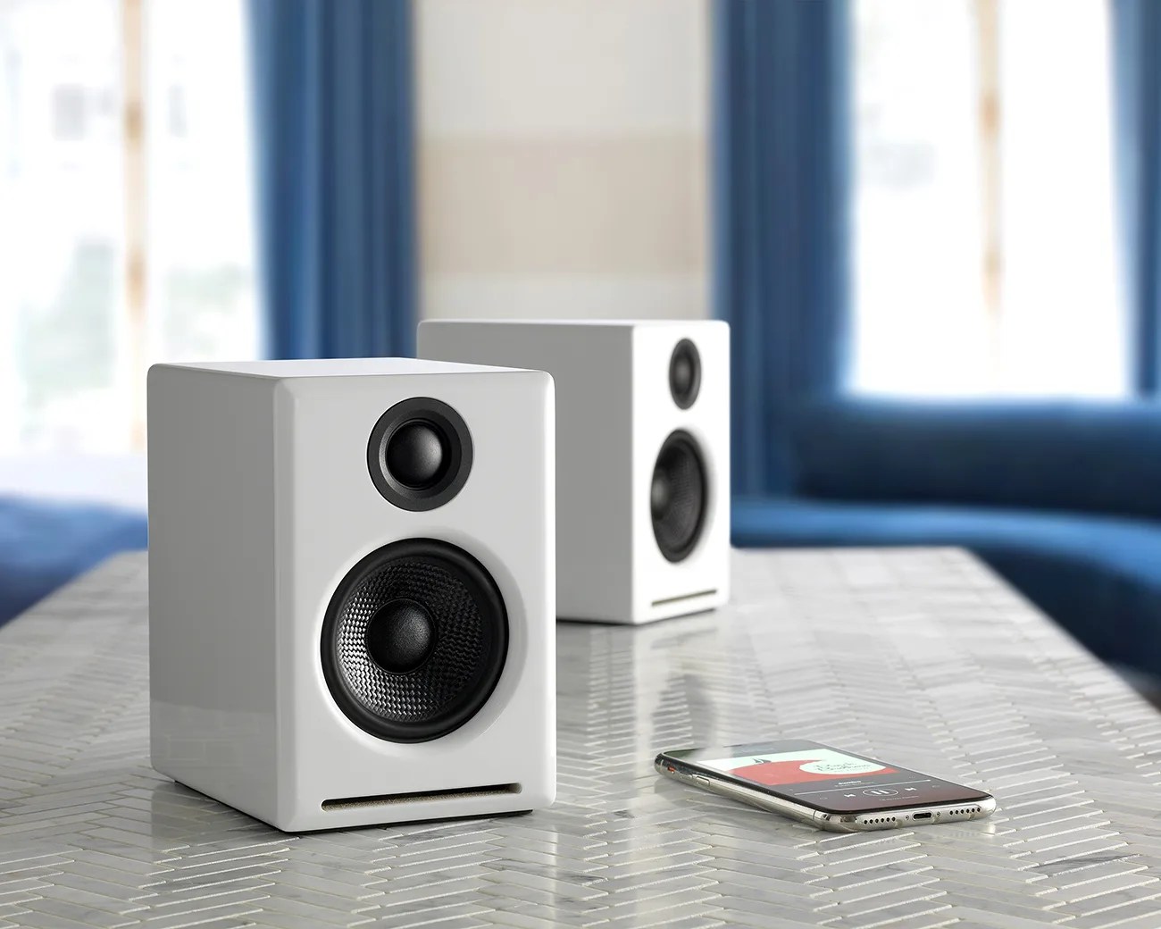 Pair of white bookshelf speakers with black drivers on a reflective table next to a smartphone playing music.