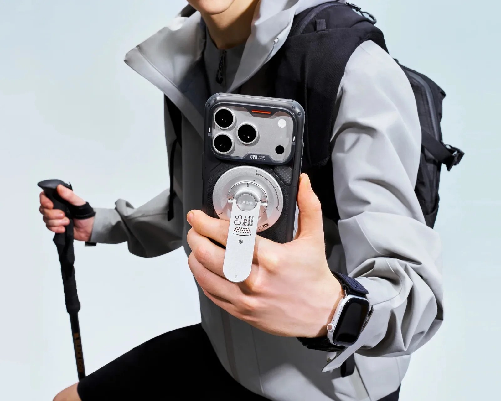 Black smartphone case with silver circular cooling attachment held by a person wearing a gray jacket and black backpack.